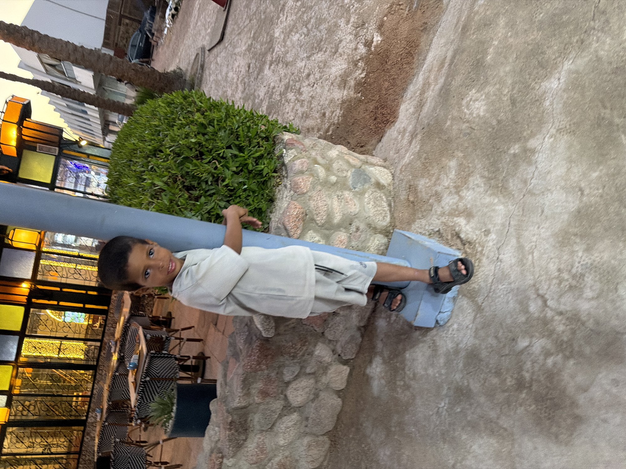 A young boy, approximately five years old, stands next to a light blue pole in what appears to be an outdoor dining area. He is wearing a light-colored short-sleeved t-shirt and matching shorts, with black sandals. The boy is looking towards the camera and has a slight smile.
Behind him, the setting features outdoor tables and chairs with black and white patterned upholstery, set up on a paved area. A potted plant is visible to the left. Further in the background, a building with large windows illuminated by yellow and orange lights suggests a restaurant or establishment. Palm trees and a white building are visible on the right side of the image, with a dark-colored car parked in the distance. The ground is a mix of concrete and sand. The overall environment suggests a location in Sharm El Sheikh, Egypt, likely a coastal resort area given the presence of palm trees and outdoor dining. The lighting indicates it is late afternoon or early evening.