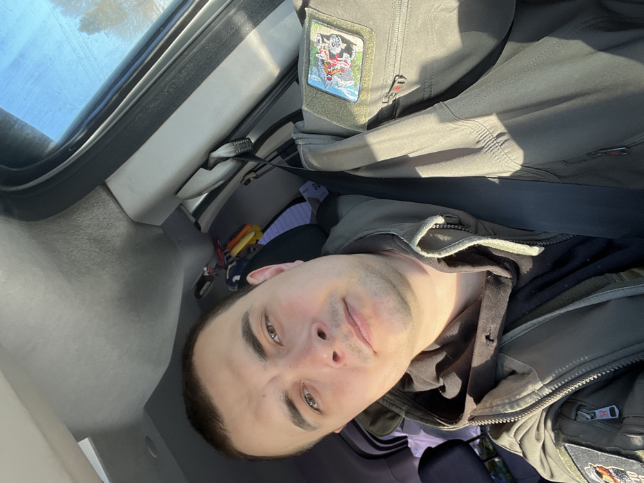 A male individual is positioned in the driver's seat of a vehicle, in what is identified as the city of Rogachovo, Russia. He has short, dark hair, brown eyes, and light stubble. He wears a black hooded garment beneath an olive-drab, tactical-style jacket with a dark, indistinct patch on the left arm. A black seatbelt is fastened diagonally across his chest. On the adjacent front passenger seat, an olive-drab tactical-style jacket or vest is draped, secured by the passenger-side seatbelt. This second garment features a rectangular patch on the right chest area depicting an off-road vehicle against a mountainous backdrop. The vehicle's interior presents a light grey headliner and pillar trim. Bright sunlight enters through the passenger window, revealing a clear blue sky and distant, bare trees outside, and creating prominent shadows within the cabin. Minor accessories are visible hanging behind the driver's headrest.