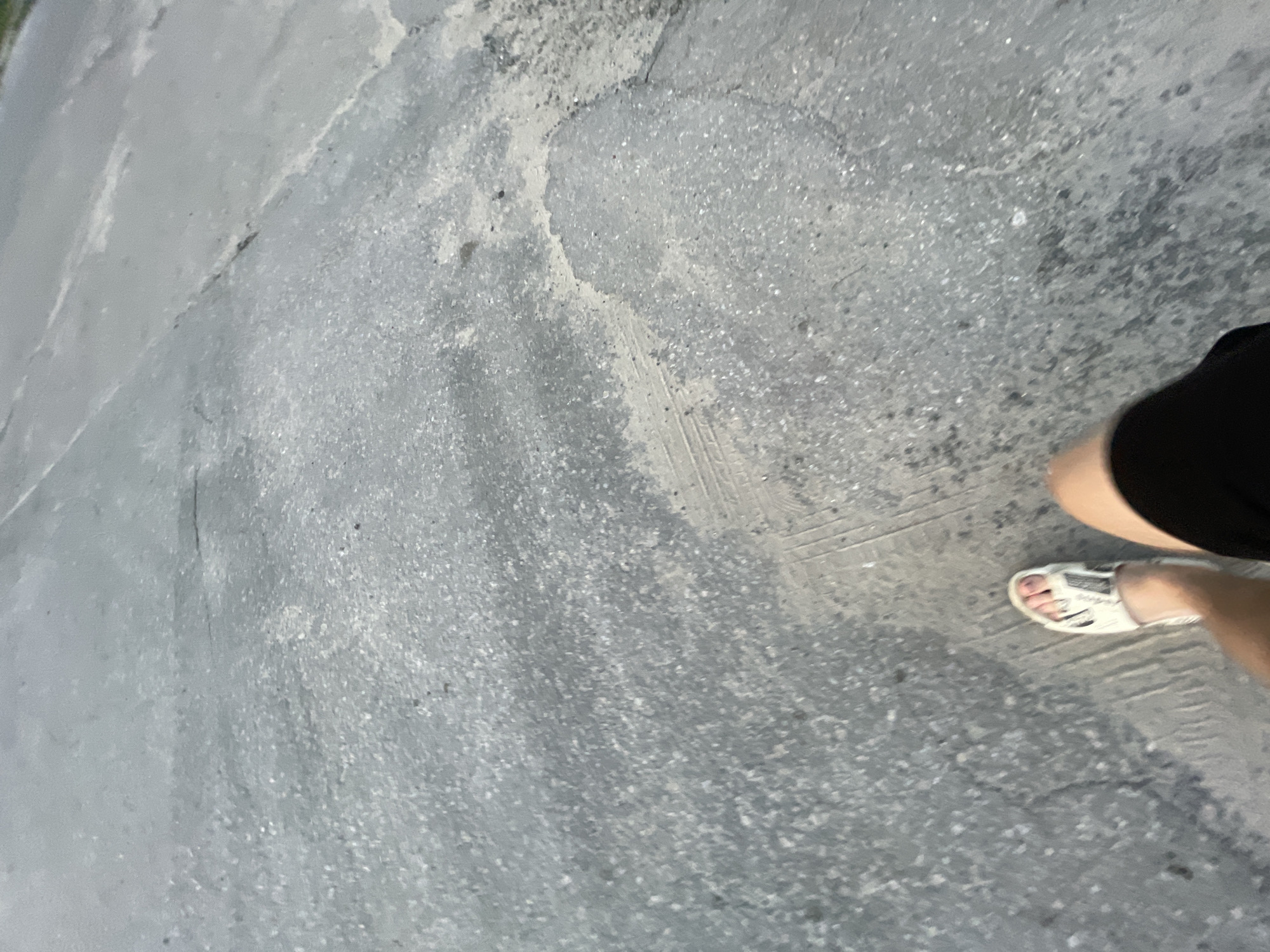 The image captures a first-person perspective of a person walking on an asphalt road in Krasnye Baki, Russia. The road surface is cracked and worn, with visible patches of lighter-colored material and some debris. Tire tracks are faintly imprinted on the asphalt. The person is wearing white sandals and dark shorts, with their legs and feet visible in the lower portion of the frame. The lighting suggests it is either early morning or late evening, as the scene is dimly lit and the sky is not visible. The environment appears to be a public thoroughfare, possibly a street or sidewalk, with no other people or structures clearly identifiable in the frame.