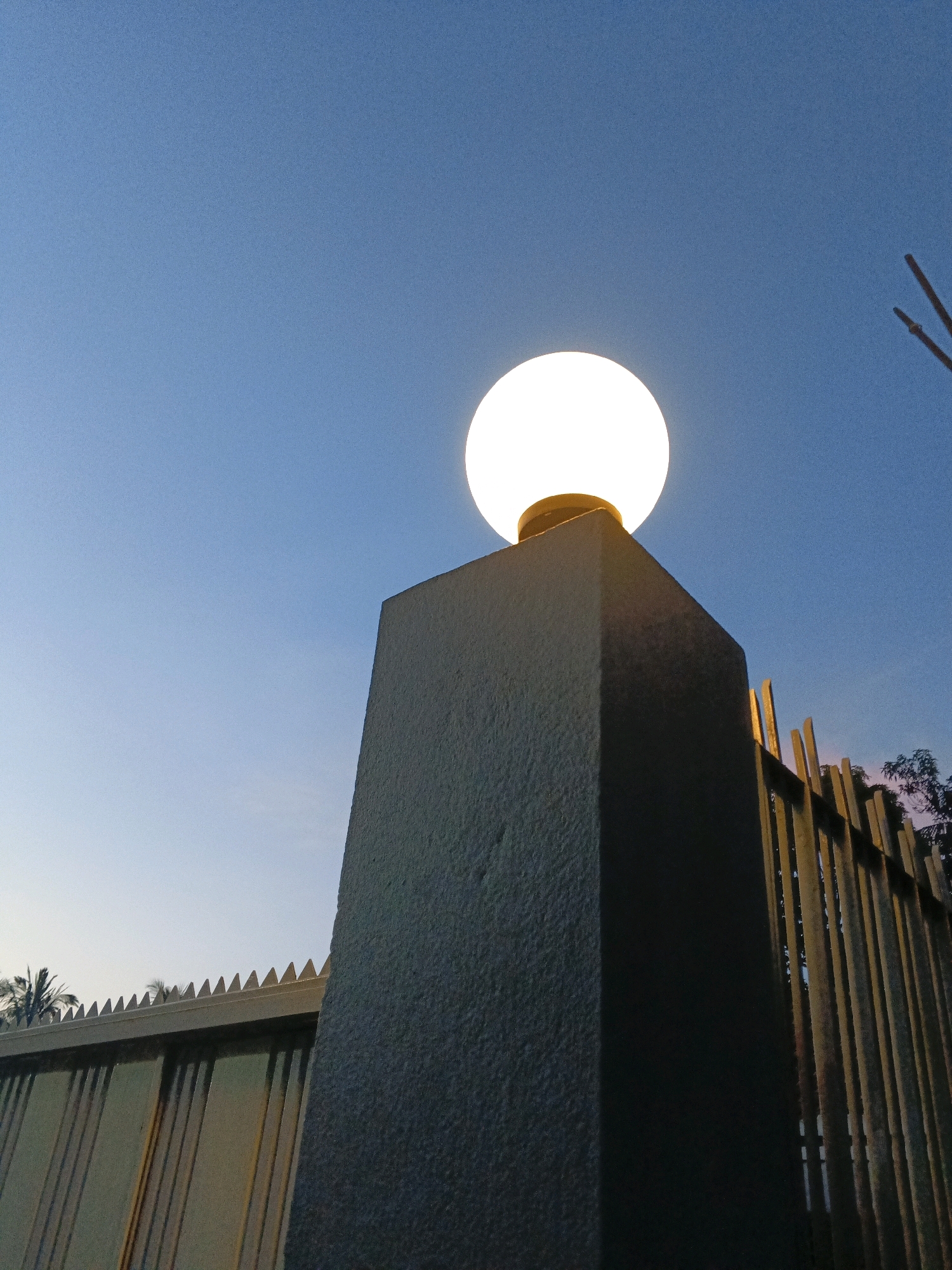 A low-angle, close-up shot captures a lit spherical lamp atop a dark grey, textured concrete pillar. The pillar is positioned in the foreground, dominating the left side of the frame. Behind the pillar, a segmented metal fence with vertical bars and a pointed, serrated top edge extends to the right. The fence is illuminated from behind by warm light, suggesting it's either sunset or sunrise, or artificial lighting within a property. To the left of the pillar, a lower fence with a similar serrated top is visible, with hints of palm trees behind it. The background is a clear, gradient blue sky, transitioning from a lighter hue near the horizon to a deeper blue overhead. The overall scene suggests an exterior location, possibly at an entrance or boundary of a property, in Sylhet, Bangladesh, during twilight. No people are present in the image.