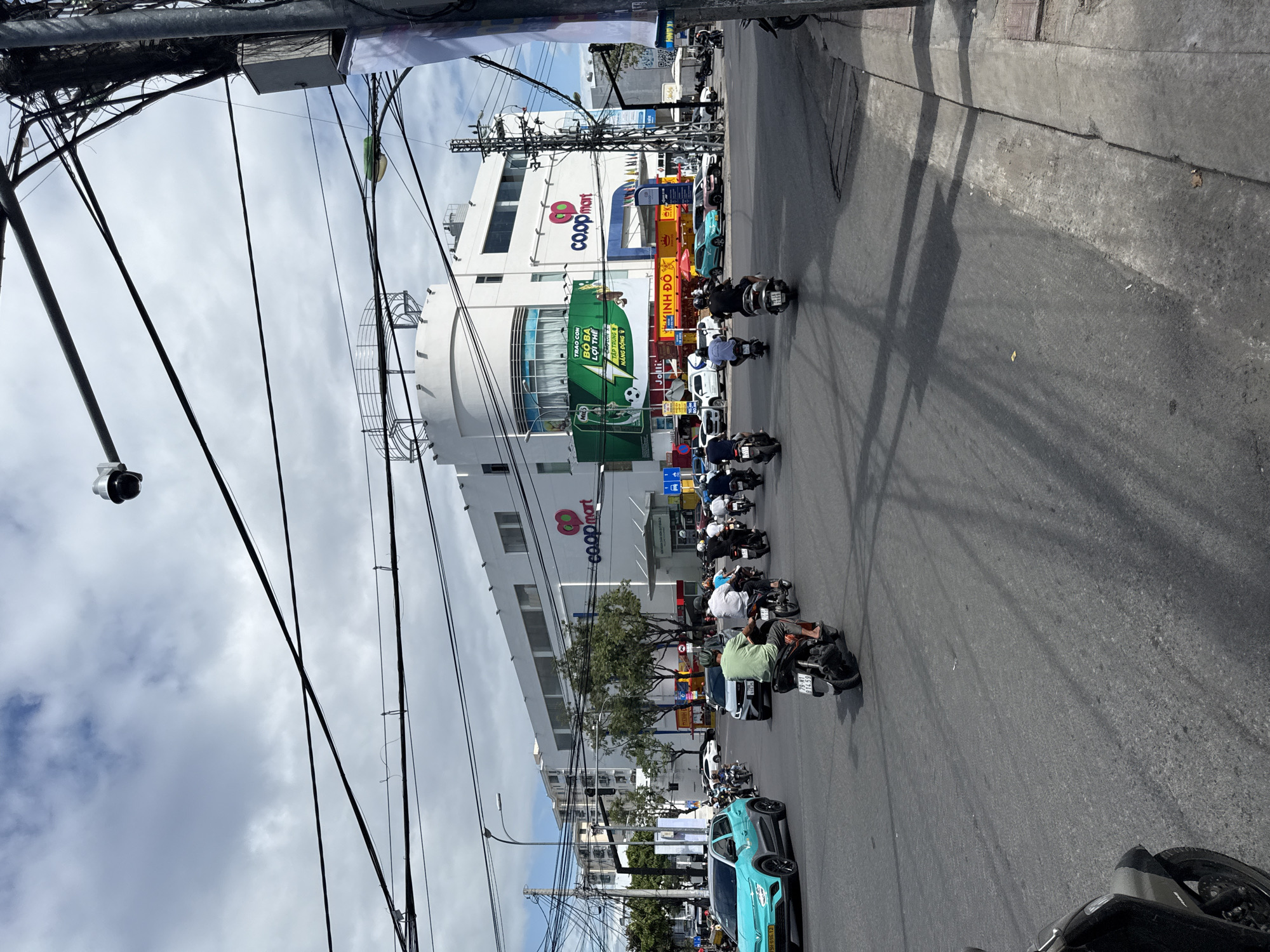 The image depicts a street scene in Nha Trang, Vietnam, dominated by a large, white Coopmart supermarket. The Coopmart building is modern, with a curved facade and prominent signage. A large green advertisement banner is displayed on the building. The street is bustling with traffic, primarily motorbikes, with a few cars visible. People on motorbikes are moving in various directions, indicating active transportation. The environment is urban, with overhead power lines crisscrossing the sky and a traffic camera mounted on a pole. The weather appears to be partly cloudy, casting shadows on the road. A turquoise car is parked on the left side of the street, and a black motorbike is partially visible in the foreground on the bottom left. The scene captures a typical day in a Vietnamese city, with its characteristic traffic patterns and commercial activity.