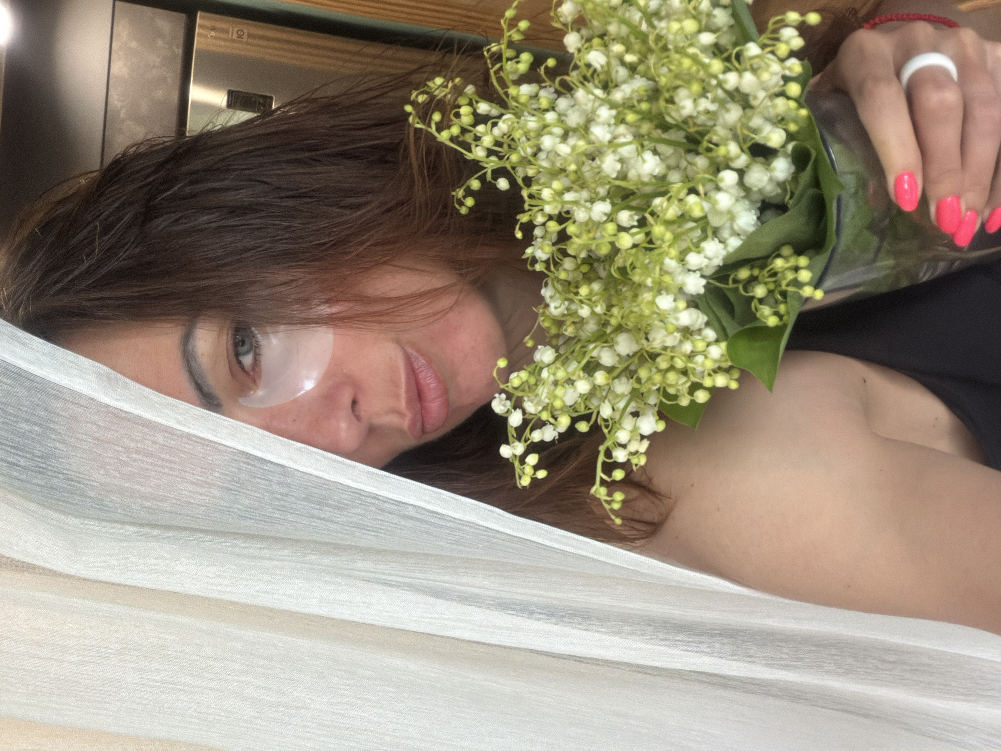 A woman with an eye mask on is lying down, holding a bouquet of lilies of the valley. She has dark hair and pink nail polish. She is covered with a white fabric. The location is likely indoors. There is no evidence of Birobidzhan, Russia.