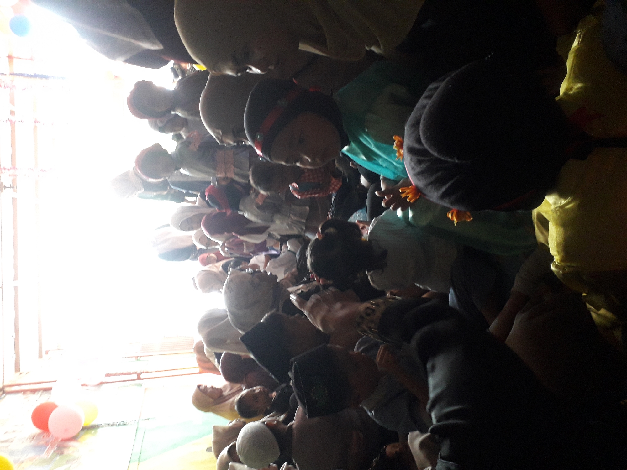 The image shows a gathering of people, primarily children and women, seated indoors. The lighting is bright and diffused, suggesting a well-lit interior space possibly with windows or an open entrance. Several individuals are wearing headscarves. In the foreground, a child in a green garment is visible, with a dark object in front of them. In the background, more people are gathered, and there appear to be some balloons and decorative elements, possibly indicating a celebration or event. The overall scene depicts a community gathering.