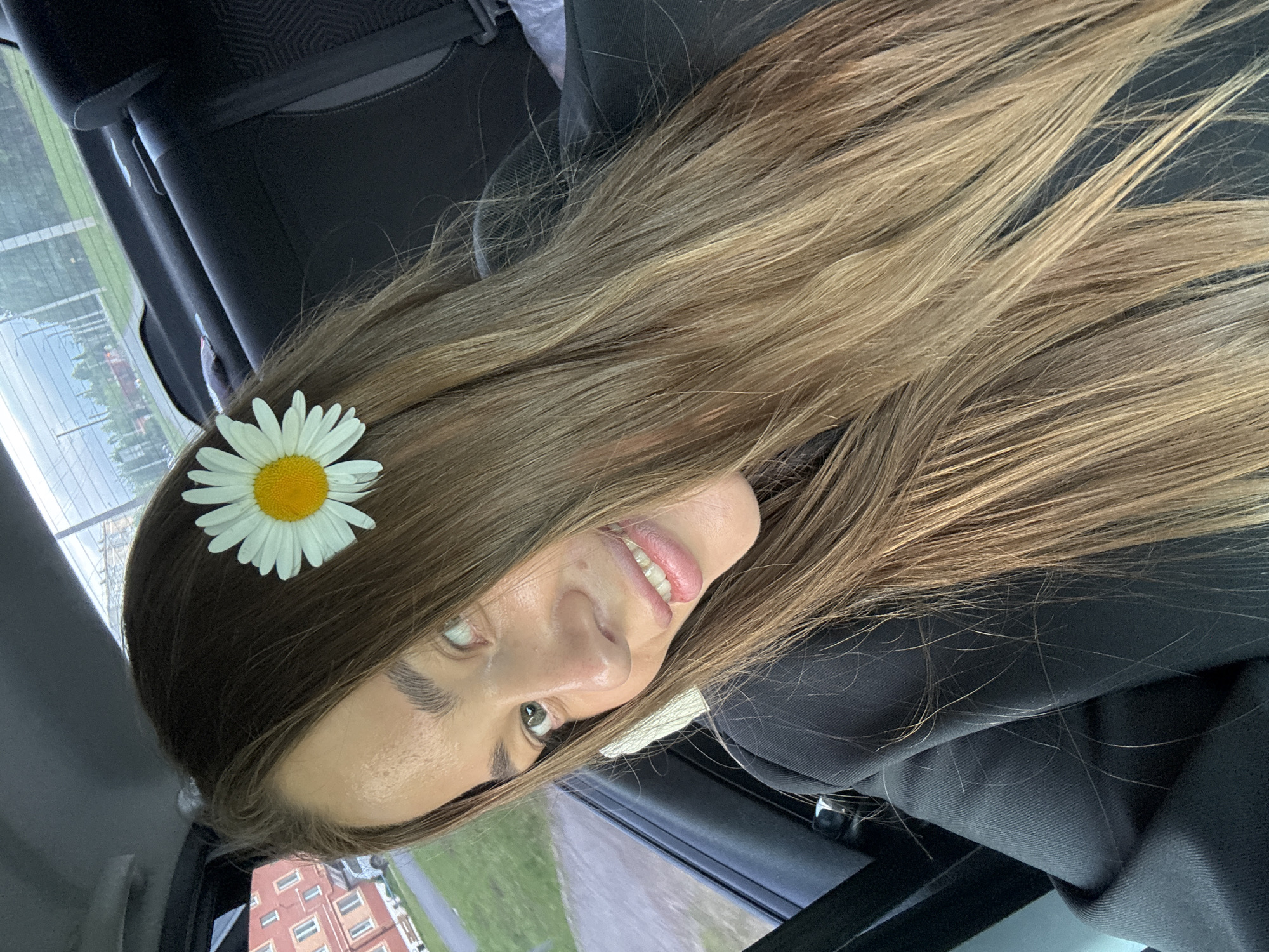 In the city of Chernushka, Russia, a young woman with long, blonde-brown hair is captured in a selfie taken from the backseat of a car. A single daisy is tucked behind her ear. The woman is wearing a black jacket, and her face is turned towards the camera, with a wide smile revealing her teeth. The car's interior is visible, including a dark gray seat. Outside the car, a building with red brick and multiple windows can be seen, along with a grassy area and a road. The background suggests an overcast day.
