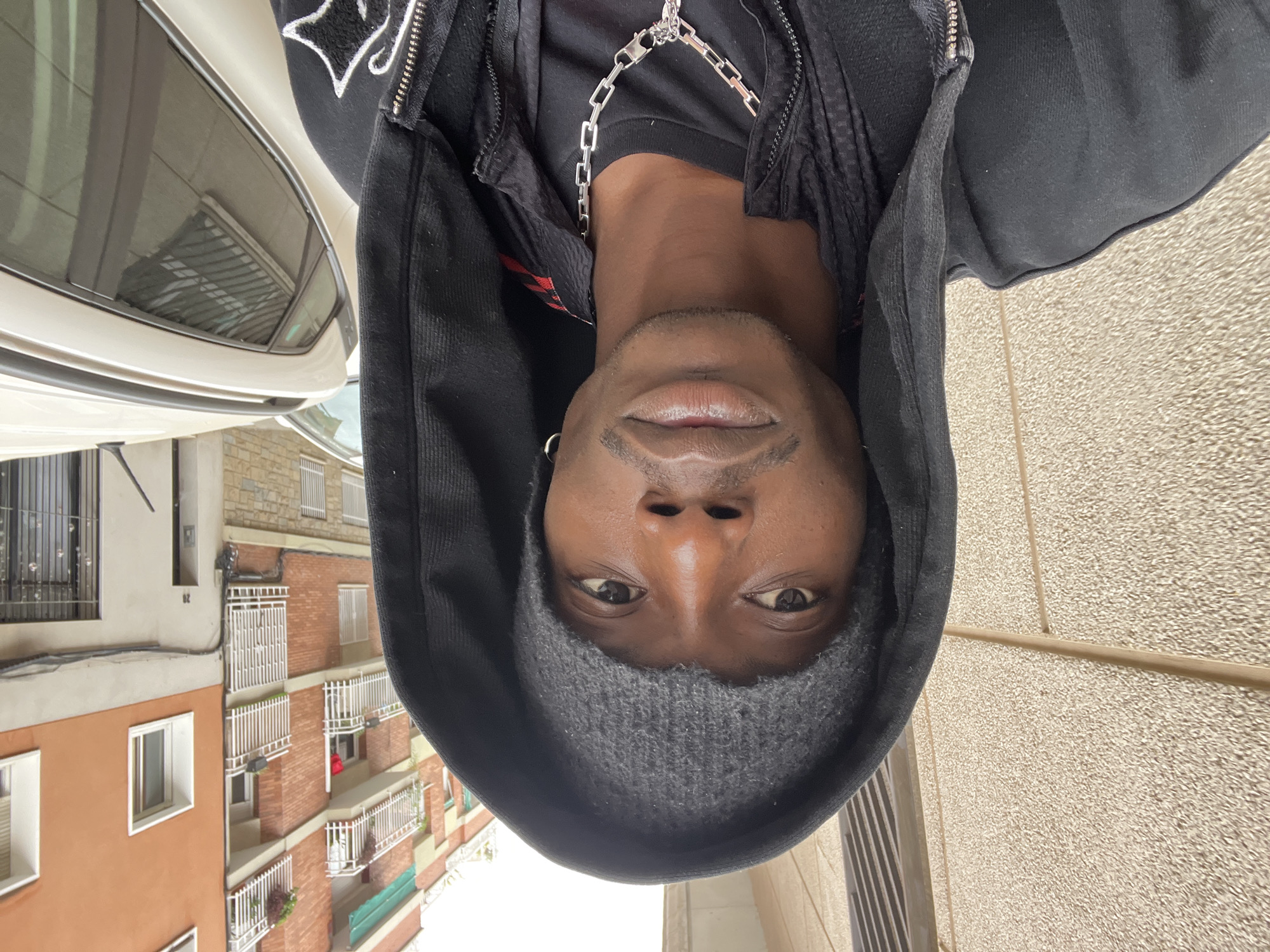A man is centered in a portrait style shot. He is wearing a black hoodie with the hood up, a dark gray beanie, and a silver chain necklace. He has dark skin, brown eyes, and closely cropped hair. The background is a city street with buildings of red brick and white balconies visible. A car is partially visible on the left side of the frame. The photograph was most likely taken in Barcelona, Spain.