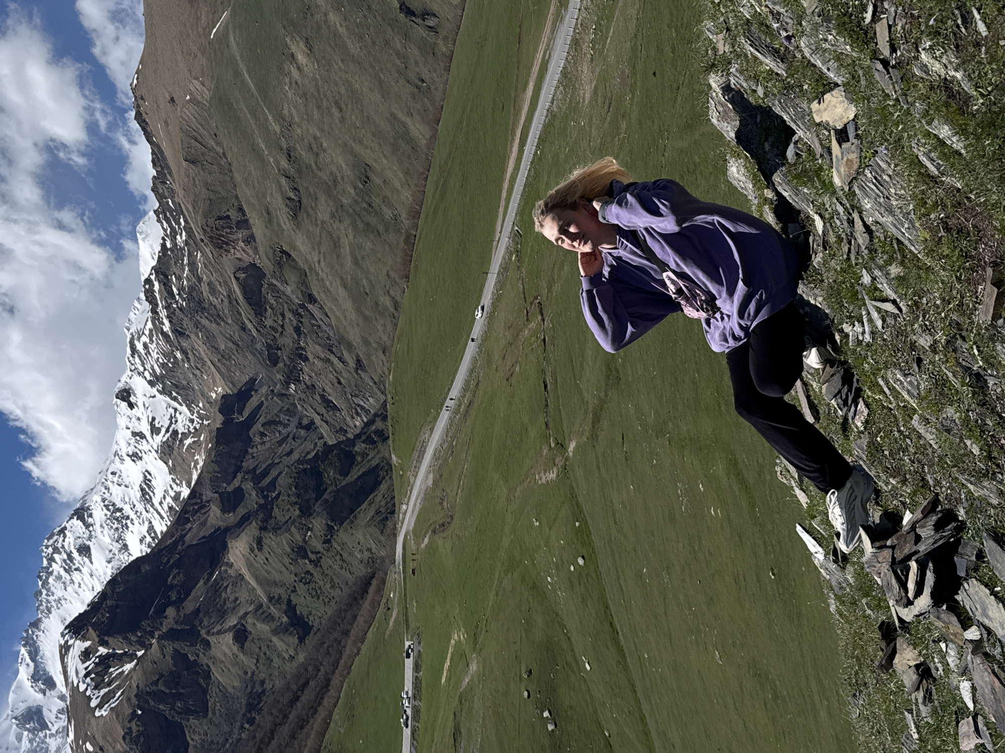A woman in a purple hoodie and black leggings stands on a rocky cliff edge. She has blonde hair and her hands are near her face. The landscape includes a green mountainside, a road with vehicles, and a snow-capped mountain range in the background. The scene appears to be taken at a high altitude in the Stepantsminda, Georgia area.