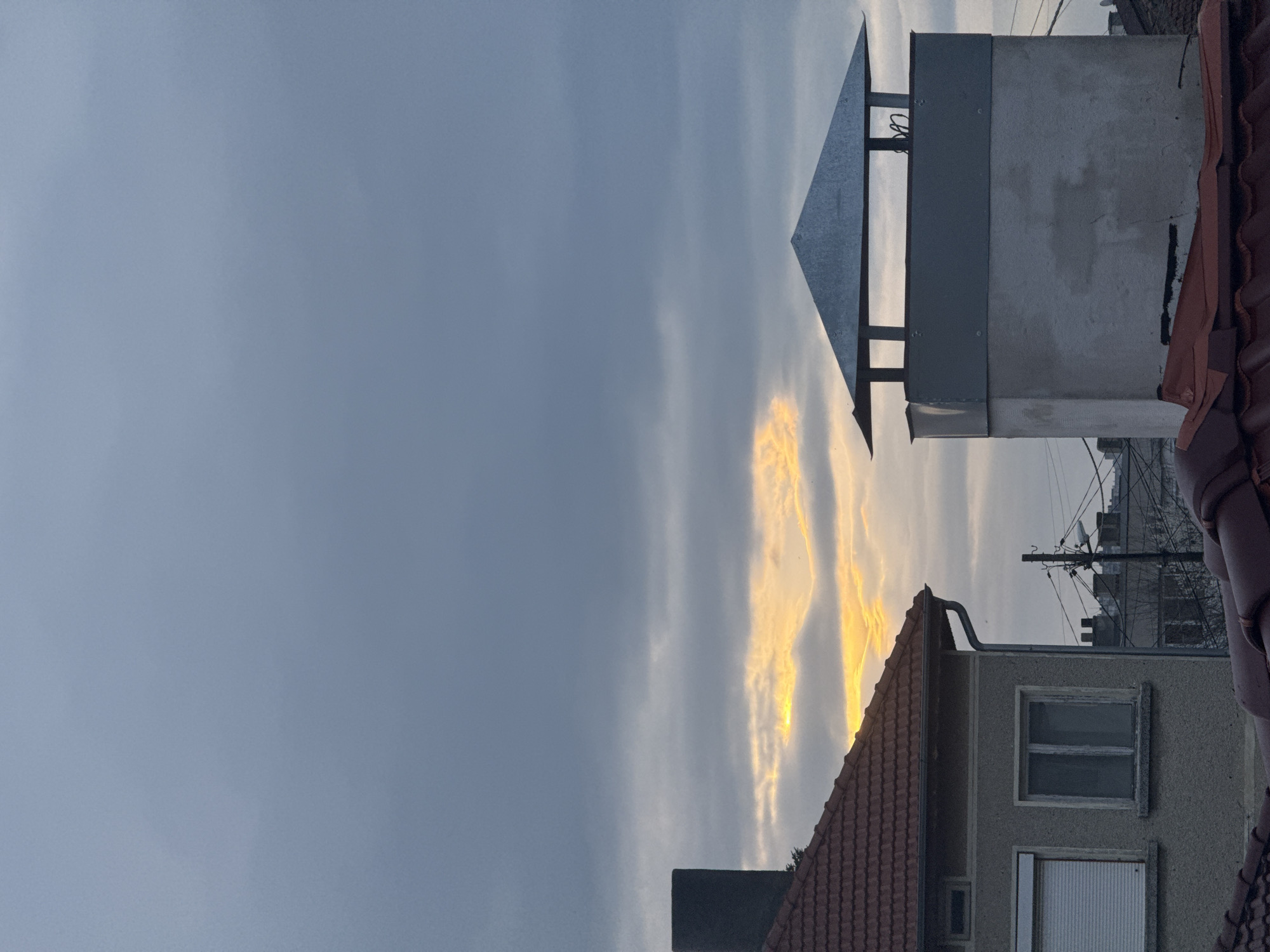The photograph, taken outdoors, features a cloudy sky with golden light streaks breaking through. In the foreground, there are rooftops and parts of buildings. A chimney with a metal cap sits atop one of the buildings. Other buildings and utility poles are visible in the background. The location is Dobrich, Bulgaria, based on visual elements typical of urban settings in the area. No people are visible in the image.