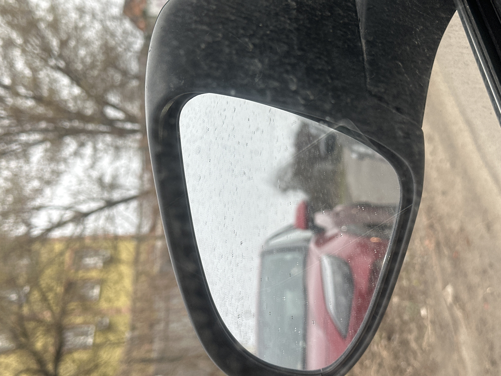 The image shows a side view mirror of a vehicle, likely a car, reflecting another vehicle, which appears to be a red truck. The mirror is covered in water droplets, and a crack is visible on its surface. Outside the mirror, a blurred building and bare trees are visible. Based on the prompt, the location is Kyiv, Ukraine.