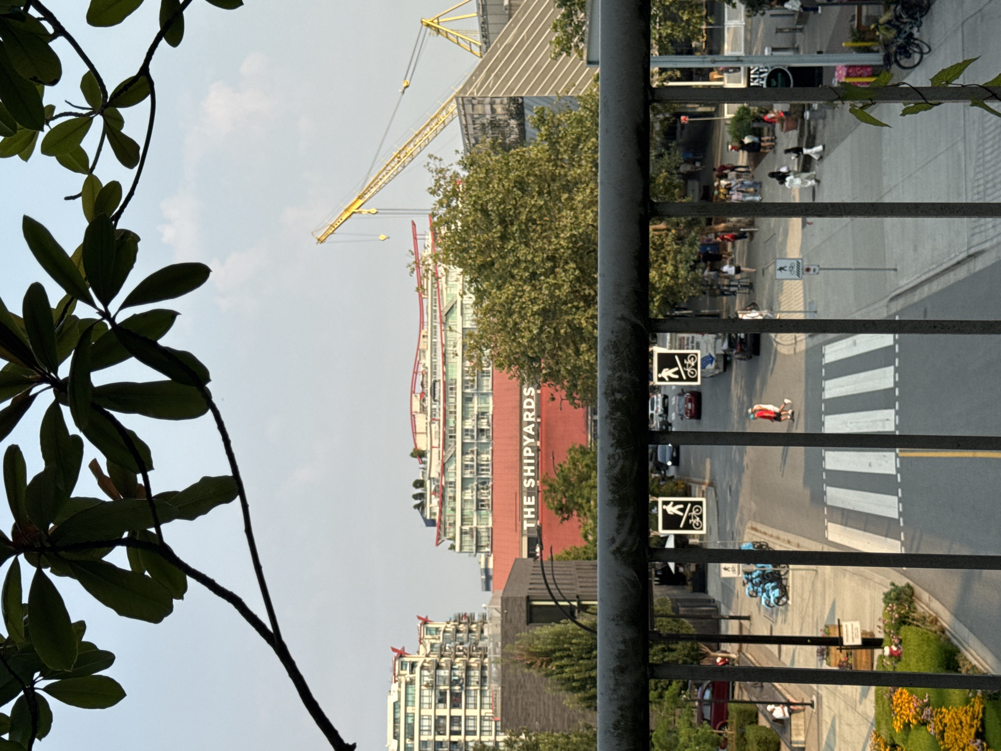 The image captures a view of Lonsdale Quay in North Vancouver, Canada, from an elevated vantage point. In the foreground, a dark metal railing with vertical bars runs across the bottom of the frame, partially obscuring the scene below. Lush green foliage hangs into the frame from the top left, silhouetted against a hazy, pale blue sky.
Beyond the railing, a street scene unfolds. Pedestrians are visible walking on the sidewalk and crossing a crosswalk. Several vehicles, including cars and blue electric scooters, are parked or driving on the street. Buildings line the street, with "THE SHIPYARDS" prominently displayed in large white lettering on a red structure in the middle ground. Behind this, taller modern apartment buildings with balconies are visible, and a yellow construction crane stands tall against the sky. The overall environment suggests an urban setting with mixed-use development, including residential and commercial spaces, along with ongoing construction. The lighting indicates it is daytime, likely late afternoon or early evening given the angle of the sun and the hazy sky.