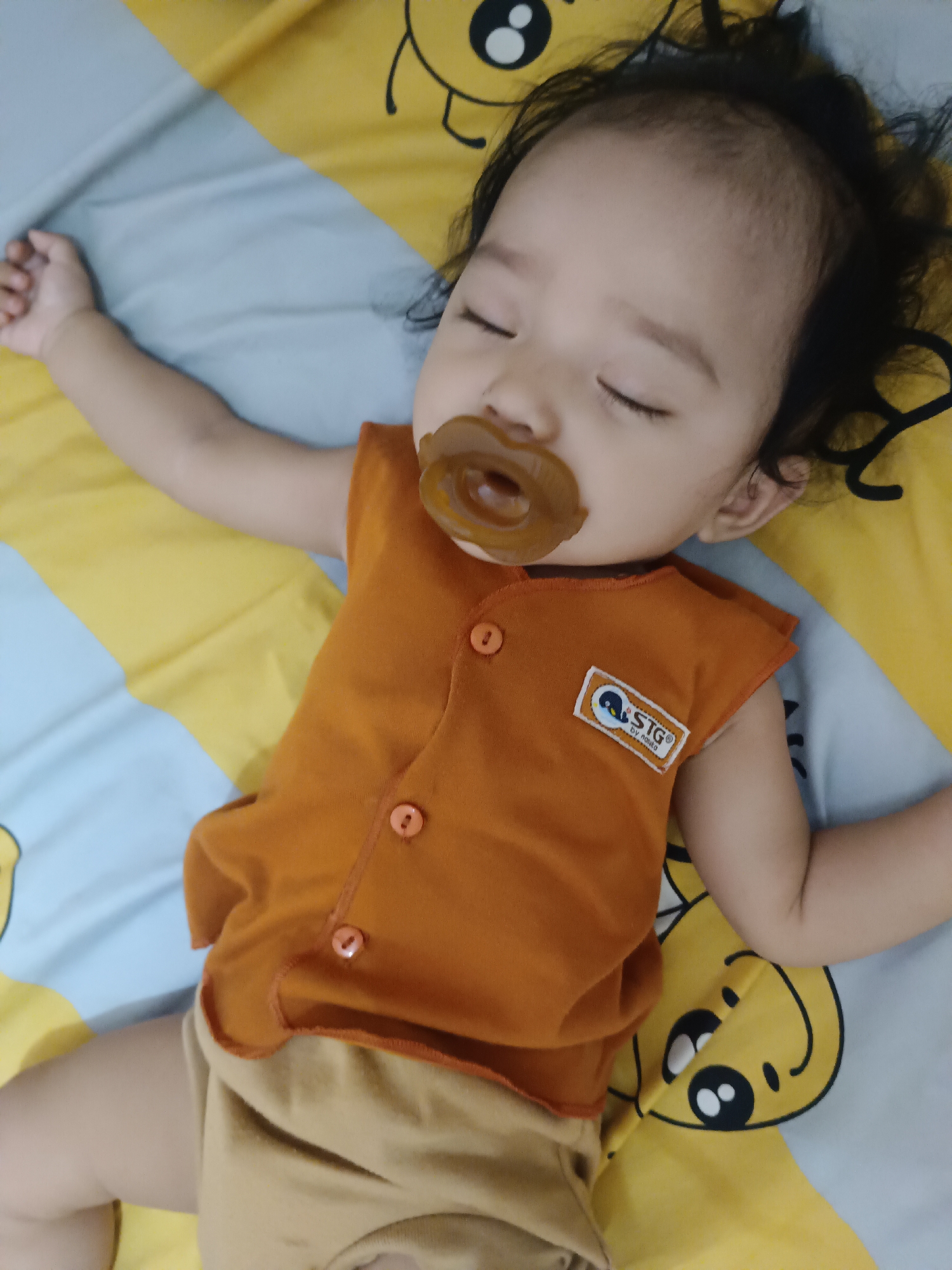 A baby is sleeping on a yellow and blue striped blanket with cartoon characters. The baby is wearing an orange sleeveless shirt and tan shorts. The baby has a pacifier in their mouth. The image is a close-up shot and does not provide enough context to determine the location in the city of Kawua, Indonesia.