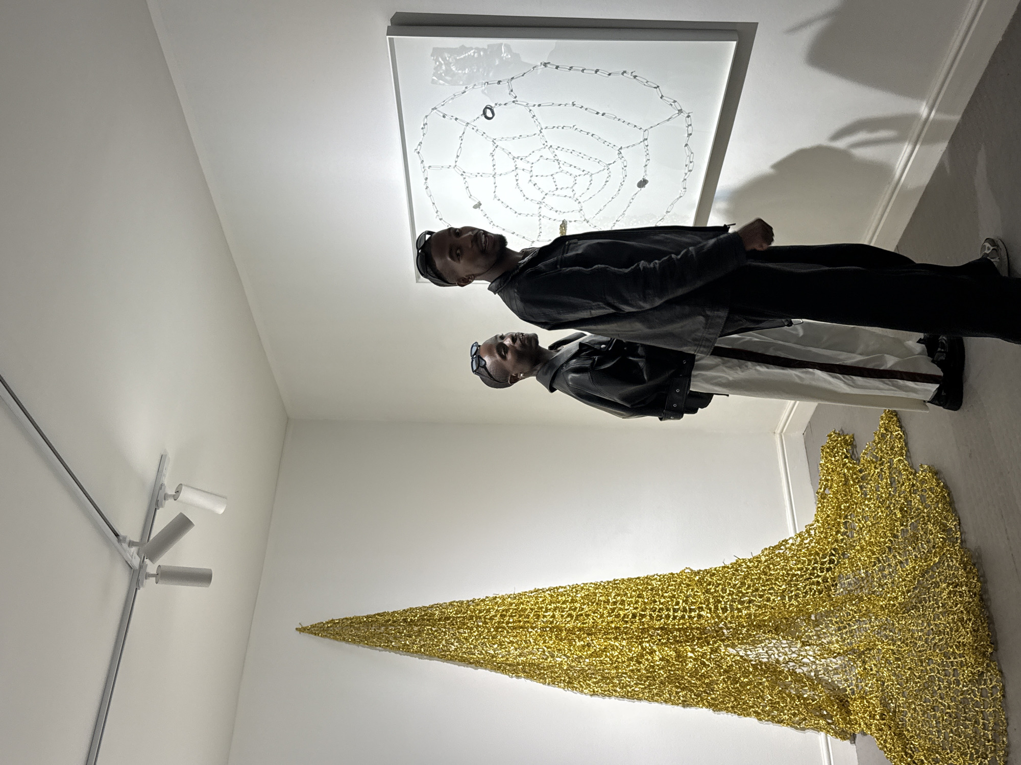 Two men stand in an art gallery in Unknown, Unknown. The man in the foreground is wearing a black leather jacket, black pants, and black shoes with white accents. The man in the background is wearing a similar outfit but with white pants with a black stripe down the side. Behind them are two pieces of art. The first is a white square with a design made of chains inside. The second is a large golden, net-like structure that is mounted on the wall. There is a track lighting system on the ceiling to the left.