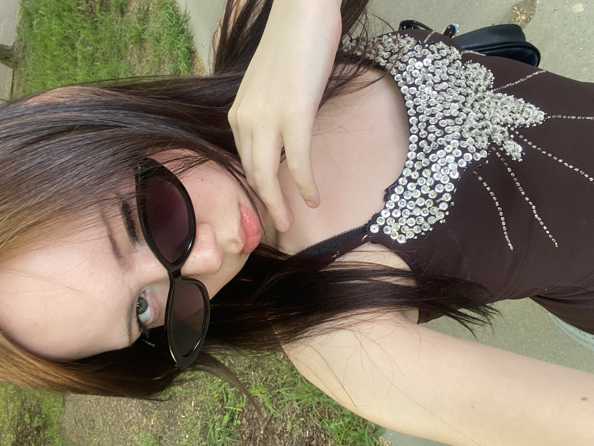 A young person is lying on their side on a patch of grass and concrete in an unknown city. The person has long, dark brown hair with blonde highlights and is wearing black sunglasses. They are wearing a brown top with silver sequin embellishments around the neckline. The person's left hand is resting on their chest. A black purse is visible in the background.