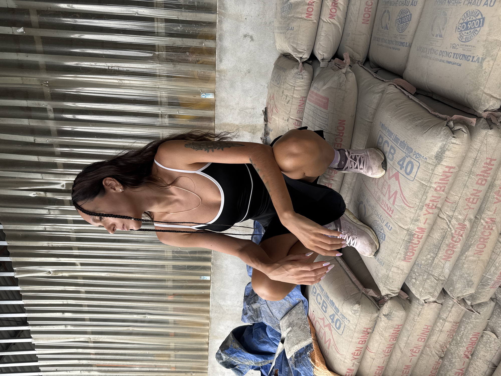 The image depicts a woman crouching near a stack of cement bags in what appears to be a warehouse or storage area in Trung Tam Ward, Vietnam. The cement bags are stacked high and labeled with "NORCEM - YÊN BÌNH" and "PCB 40." The woman has braided hair, a tank top and black shorts. She wears sneakers and has a tattoo on her arm.
The environment has metal walls. Part of a tarp is also visible.