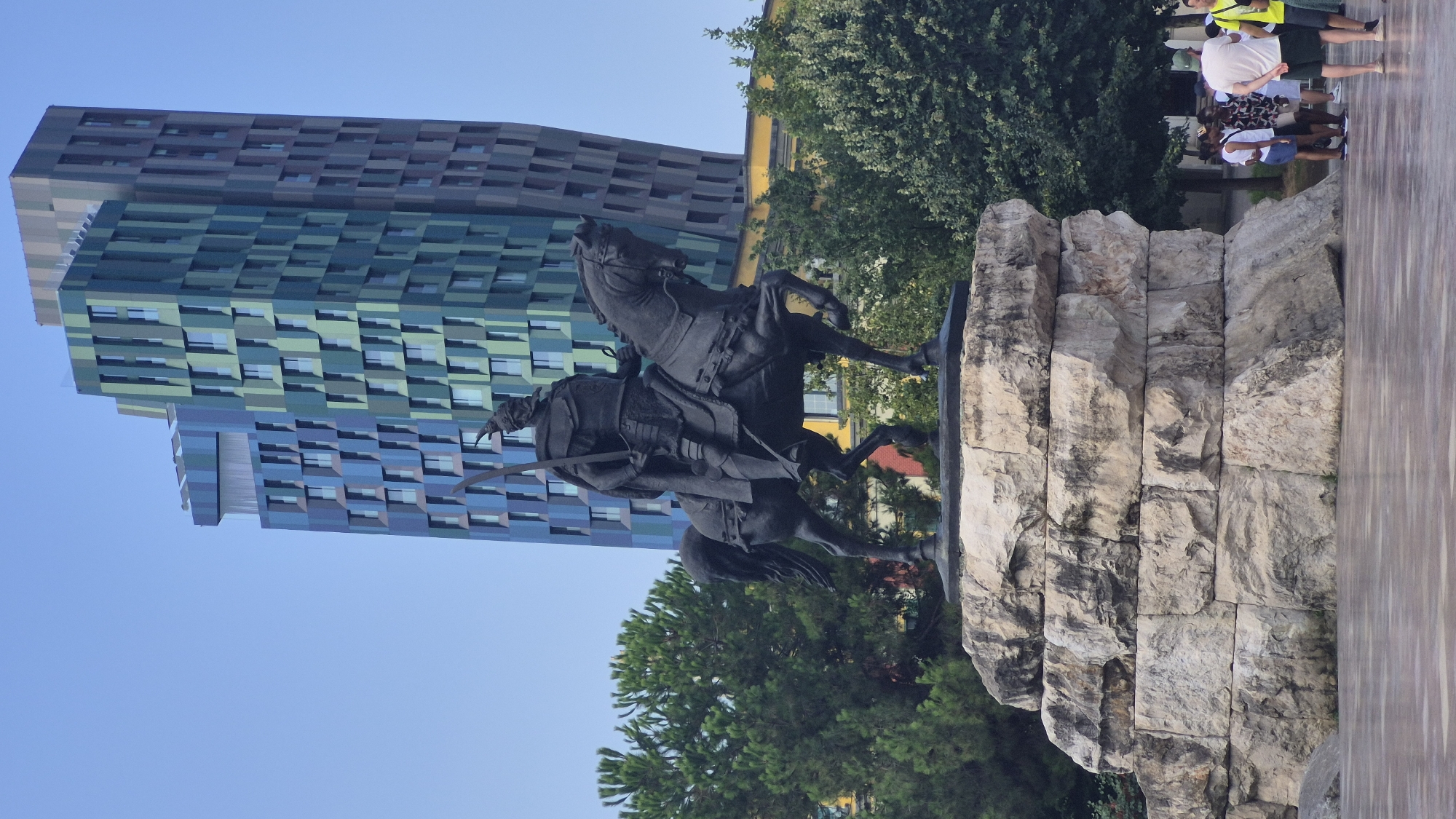 The image captures Skanderbeg Square in Tirana, Albania, a prominent public space dominated by a large equestrian statue of the Albanian national hero, Gjergj Kastrioti Skanderbeg. The statue, cast in dark metal, depicts Skanderbeg in armor, holding a sword aloft, mounted on a powerful horse in mid-stride. It rests on a substantial, multi-tiered stone pedestal.
Behind the statue, the cityscape of Tirana is visible. A striking modern building with a distinctive facade of blue and green panels, the Tirana Times Square, rises into the clear blue sky. This architectural landmark contrasts with the more traditional architecture suggested by a yellow building partially visible to the right of the modern structure. Lush green trees, likely pines and other deciduous varieties, surround the statue and the square, providing a natural element against the urban backdrop.
In the foreground, several people are casually gathered on the paved ground of the square. They appear to be tourists or locals, some dressed in summer attire, with one individual wearing a white t-shirt and dark shorts and another in a yellow t-shirt and dark shorts. Their presence indicates a public space actively being utilized. The paving stones in the immediate foreground are wet, suggesting recent rain or cleaning. The overall impression is one of a historical monument set within a vibrant, modern urban environment.