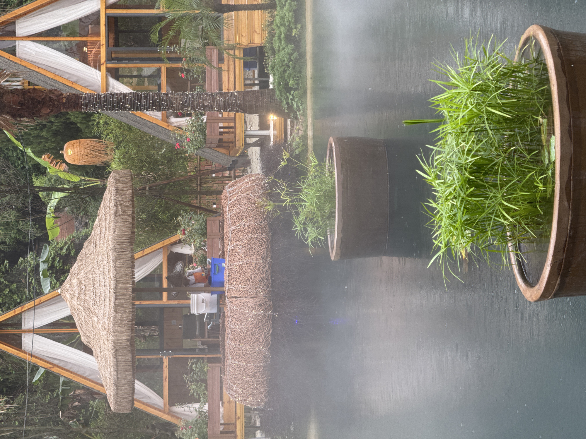 The image shows an outdoor scene, possibly in Guilin, China, given the architectural style and vegetation. It's raining. A small thatched-roof structure sits near what appears to be a pond or wet surface reflecting the scene. Nearby are cylindrical planters with greenery. A wooden structure with white curtains is visible in the background, suggesting a resort or relaxation area. A palm tree trunk is also visible. The foreground is dominated by the wet surface, blurring the reflection of the background elements.