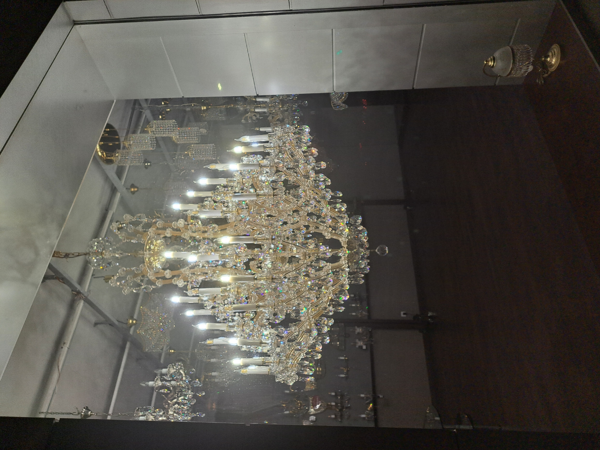 The image depicts a chandelier with multiple crystal components and numerous light bulbs, seemingly suspended in a display case or window. The composition appears to be a reflection, with the left side showing the interior of what seems to be a retail space or showroom, reflected on a glass surface, while the right side reflects a darker, possibly interior space. Other light fixtures are visible on the ceiling in the reflection. The image was taken indoors, and there is no clear indication of its location in Yerevan, Armenia, or any people present. The focus is on the chandelier and the interplay of light and reflections.