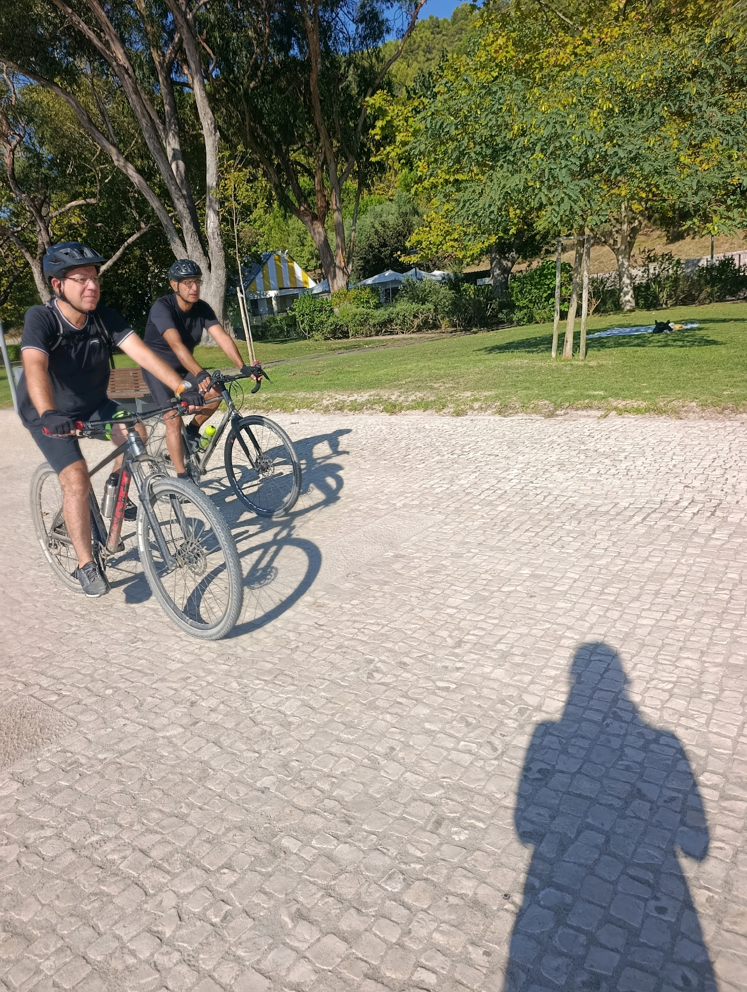 Two men in black t-shirts, black shorts, helmets, and gloves are cycling on a cobblestone path. The man in the foreground, on the left, is wearing glasses and cycling a dark-colored bicycle with a red water bottle cage. The man behind him, on the right, is cycling a lighter-colored bicycle. In the background, there is a grassy area with trees and a yellow and white striped structure that appears to be a tent or canopy. A person is lying on a blue blanket in the grass. The sky is blue and clear. The scene is set in the Parque do Bonfim in Setúbal, Portugal, known for its cobblestone paths and recreational areas.