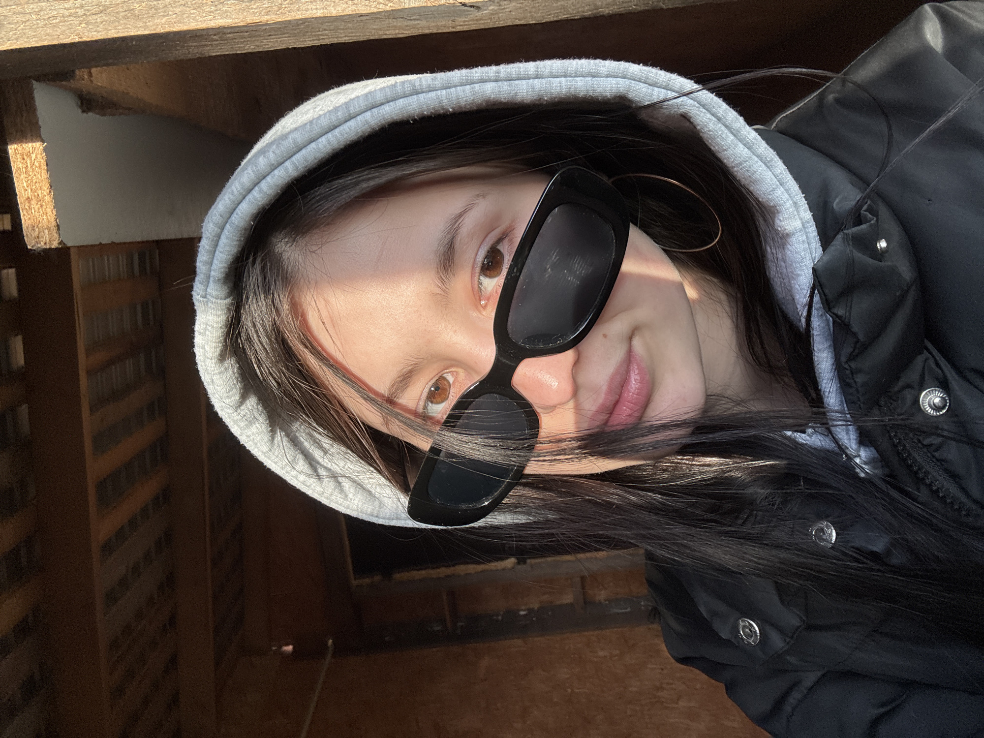 A young individual is captured in a tilted, close-up selfie, looking directly at the camera. They have dark, long hair, light skin, and brown eyes, one of which is visible over a pair of stylish black rectangular sunglasses. A subtle, natural-looking pink lipstick is worn on their lips, which curve into a slight smile or smirk.
The subject is wearing a light grey hooded sweatshirt with the hood up, underneath a black puffer jacket that features silver snap buttons. A large, golden hoop earring is visible on their right ear (viewer's left).
The background suggests an outdoor or semi-enclosed space, likely a balcony or a similar structure made of wood, with visible wooden beams and planking. Bright natural sunlight casts strong shadows, indicating it is daytime and sunny. In the distance, particularly noticeable in the cropped hints, there appears to be a multi-story building, suggesting an urban or suburban environment in or around Фоки, Russia. There is no visible text in the image.