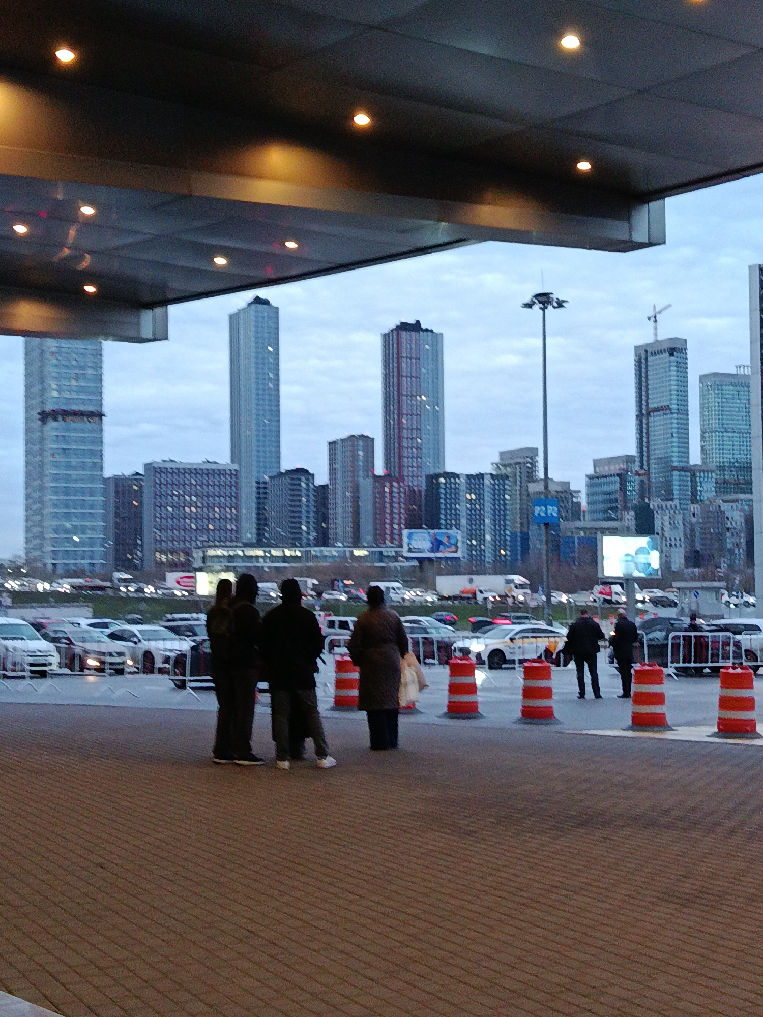 The image shows a group of people standing on a brick-paved area under a modern canopy-like structure, seemingly waiting or observing the cityscape in the background. The city skyline includes several tall buildings, some under construction, with varying architectural styles. There's a light pole and electronic billboards adding to the urban landscape. A row of cars is parked behind the people, separated by traffic cones. The sky is overcast, suggesting a cloudy day.