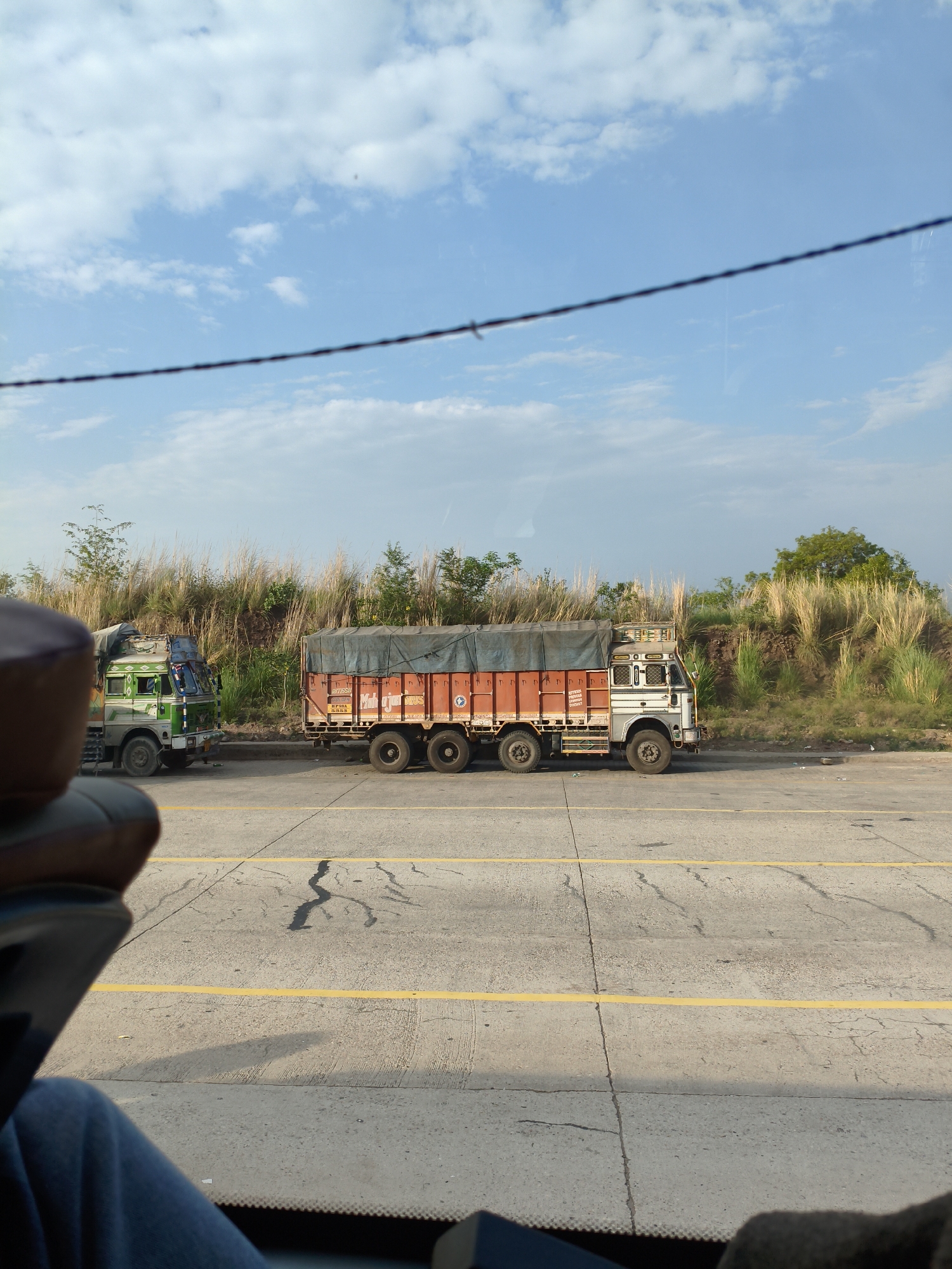 The image depicts a roadside scene with two trucks parked on the right side of a concrete road marked with yellow lines. In the foreground, a portion of a vehicle's interior is visible, including a dark brown leather seat and a person's legs covered in blue fabric. A thin, dark wire runs horizontally across the upper part of the image, partially obscuring the blue sky with scattered white clouds.
The first truck, on the left, is green and white with a loaded trailer covered by a green tarp. It has a distinctive, ornate front cab. The second truck, to its right, is larger and predominantly orange with a blue tarp covering its trailer. This truck also features signage on its side, which is partially legible. Both trucks are positioned along a raised embankment covered in tall, dry grass and scattered green foliage. The road surface appears weathered with cracks and dark stains. The overall impression is that of a sunny day on a highway in India, likely in the vicinity of Anandpur Sahib, given the regional context of similar roadside scenes.