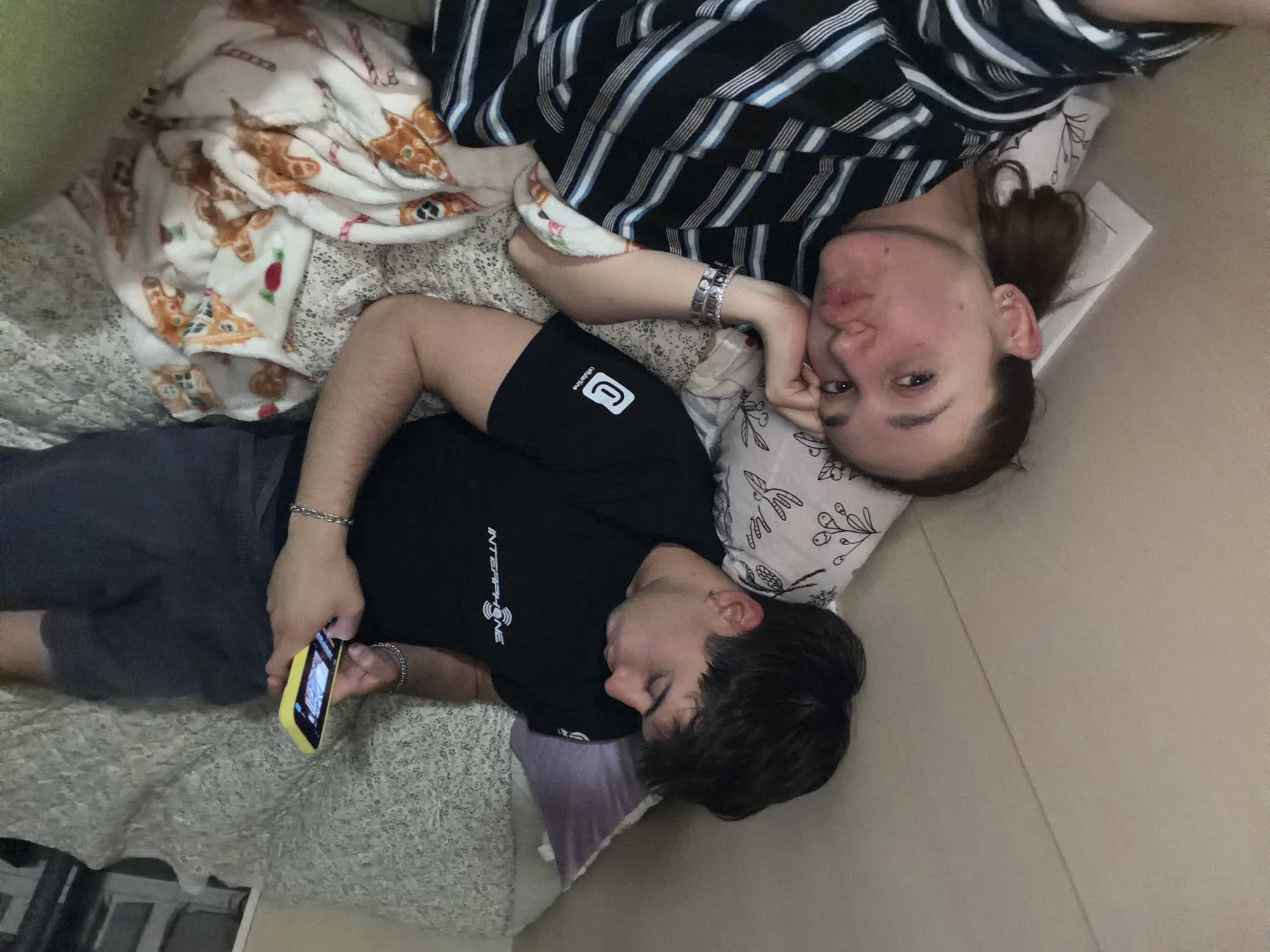 This media file captures two individuals, a man and a woman, relaxing together indoors, likely in a bedroom or living area in Sofia, Bulgaria. The image is taken from above, looking down on them as they lie on a bed or couch.
The woman is positioned in the upper right quadrant of the image. She has brown hair pulled back and is wearing a black and white striped shirt. A silver bracelet adorns her left wrist. She is looking towards the camera with a relaxed expression, her left hand resting against her face. Her head rests on a white pillow adorned with a black floral or botanical pattern.
The man is in the lower left, lying beside her. He is wearing a black t-shirt with the word "INTERPHONE" visible in white lettering, and dark shorts that feature a white "CollabMarine" logo. A silver chain bracelet is on his left wrist. He is holding a yellow-cased smartphone in his hands, seemingly engrossed in its screen. His head rests on a similar white, patterned pillow, with a purple pillow also visible beneath his neck.
A cozy atmosphere is created by the bedding, which includes a white blanket patterned with gingerbread houses and Christmas cookies, and a grey textured sheet or blanket underneath. The background shows parts of light-colored walls and possibly a window or mirror reflecting light at the bottom left. The overall scene depicts a casual moment of shared relaxation and downtime. The lighting suggests it is daytime.