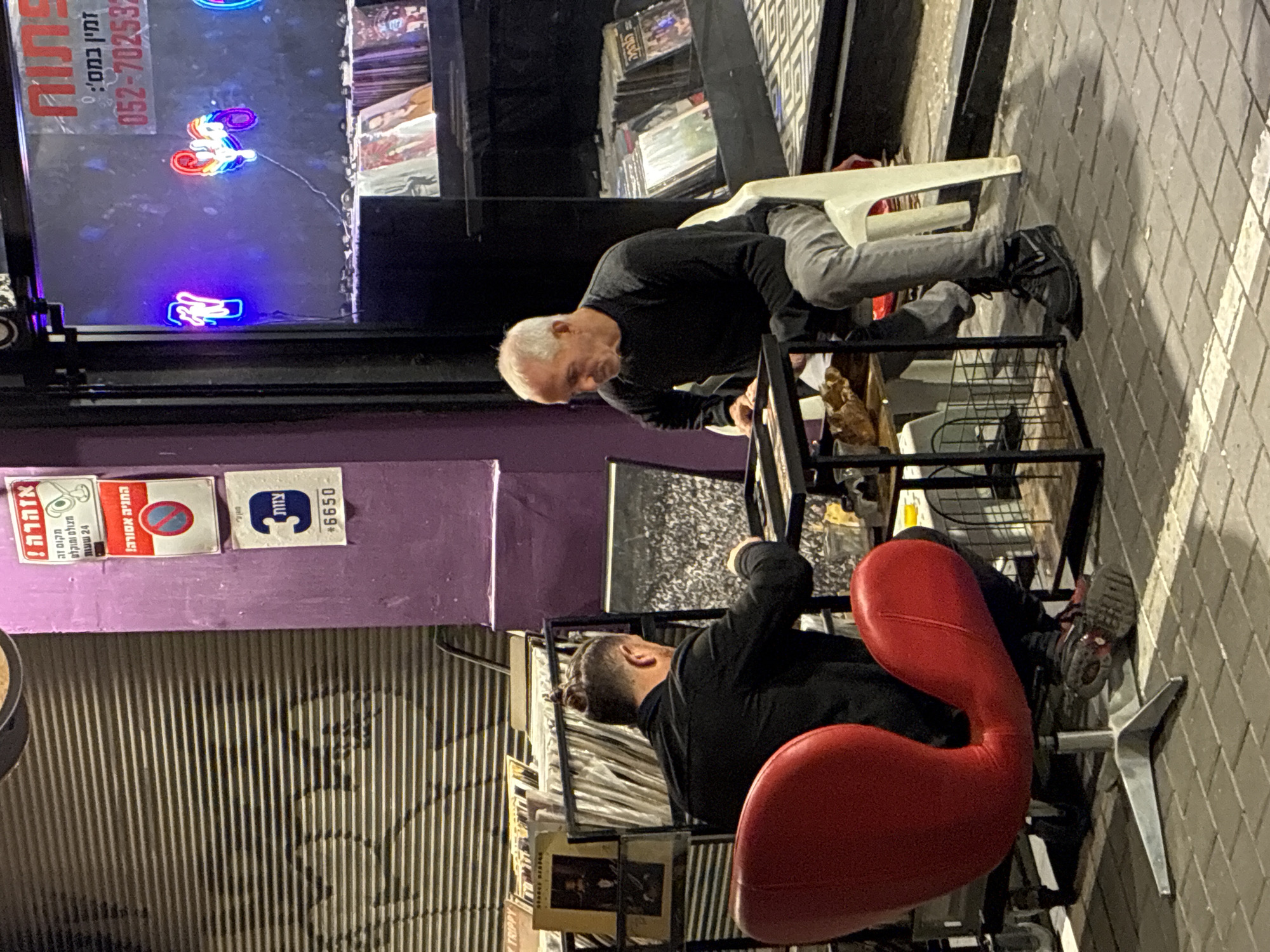 The image captures an outdoor scene in Tel-Aviv, Israel, at night, where two men are engaged in a record selection process. One man, with short grey hair, wears a black long-sleeved shirt and grey pants, as he leans over a black metal shelving unit filled with vinyl records. He sits on a white plastic chair. Opposite him, another man, with dark hair, is seated on a red swan-shaped chair, also examining the records. He's dressed in a black long-sleeved shirt.
Behind the men, a closed, silver-grey corrugated security gate is visible. A purple wall above the gate has multiple stickers, including a warning sign written in Hebrew, a "No Parking" sign, and a "3" sticker. The store's exterior window displays neon lights shaped like unicorns and a hand, with additional information in Hebrew including store name "פתוח" (Open), phone number and "זמין במס':". More record albums are stacked inside the storefront.
The background shows a dark, brick-paved sidewalk leading away into the night. The scene suggests a relaxed, informal setting, typical of an outdoor record store in the city.