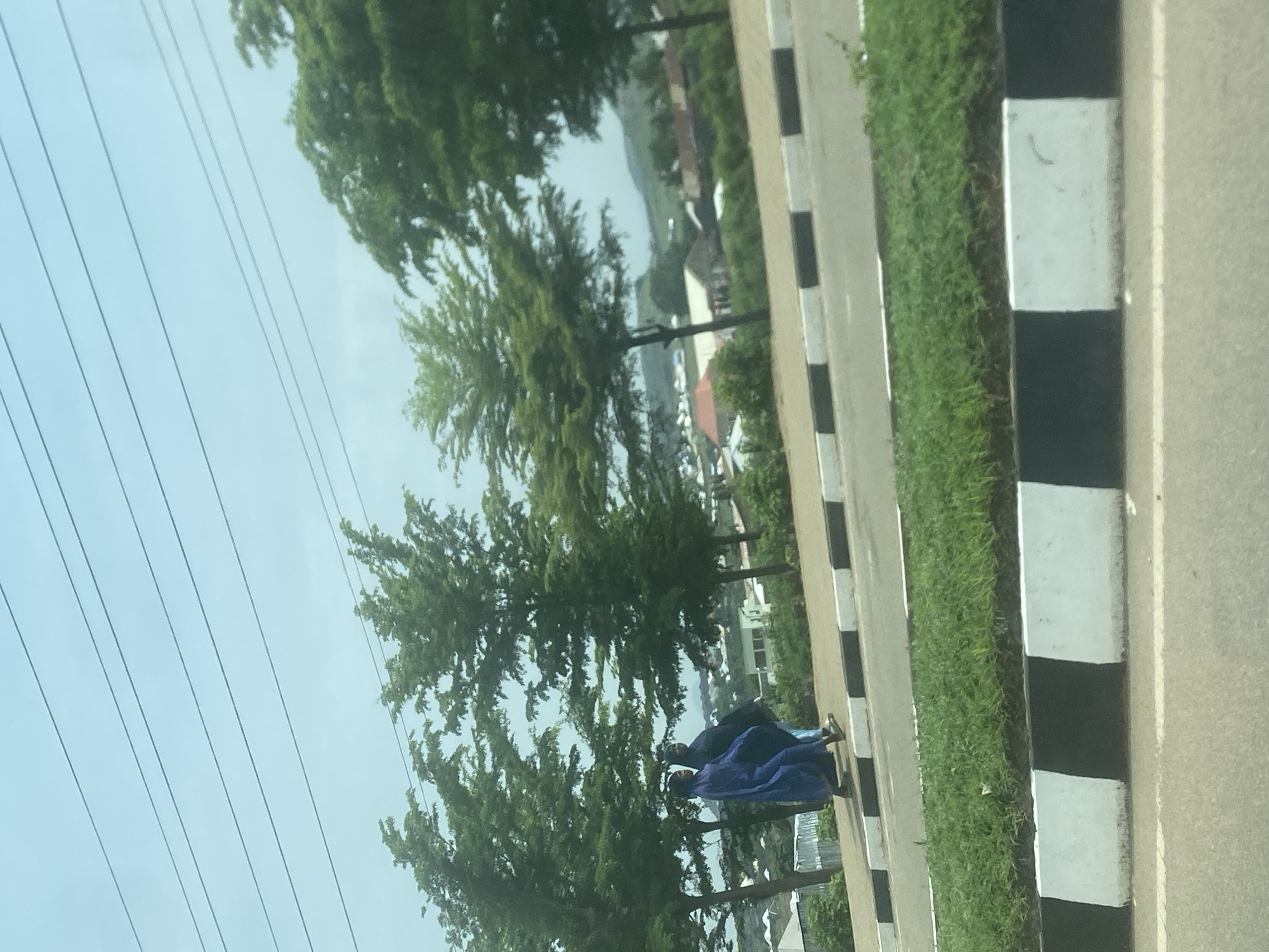 This media file depicts a roadside scene in Gosa, Nigeria, likely viewed from a vehicle. The upper portion of the image shows a light blue sky traversed by several parallel overhead power lines.
Below the sky, numerous green trees with dense foliage line the side of the road, suggesting a verdant environment. Through and beyond the trees, hints of distant buildings and possibly hills are visible, indicating a semi-urban or rural-urban fringe setting.
In the mid-ground, a paved road runs parallel to a distinct black and white striped curb or median. Adjacent to this curb is a narrow strip of green grass. Two people are seen walking along a path next to the grassy strip. One individual is dressed in a dark blue, loose-fitting garment, and the other in a lighter blue, similar attire. They appear to be walking in the same direction, with their backs partially turned to the viewer, and their faces are not clearly discernible.
The lighting suggests it is daytime, with diffuse light indicating either an overcast sky or bright conditions without direct harsh sunlight. There is no visible text present in the image. The black and white striped road markings are a prominent detail of the road infrastructure.