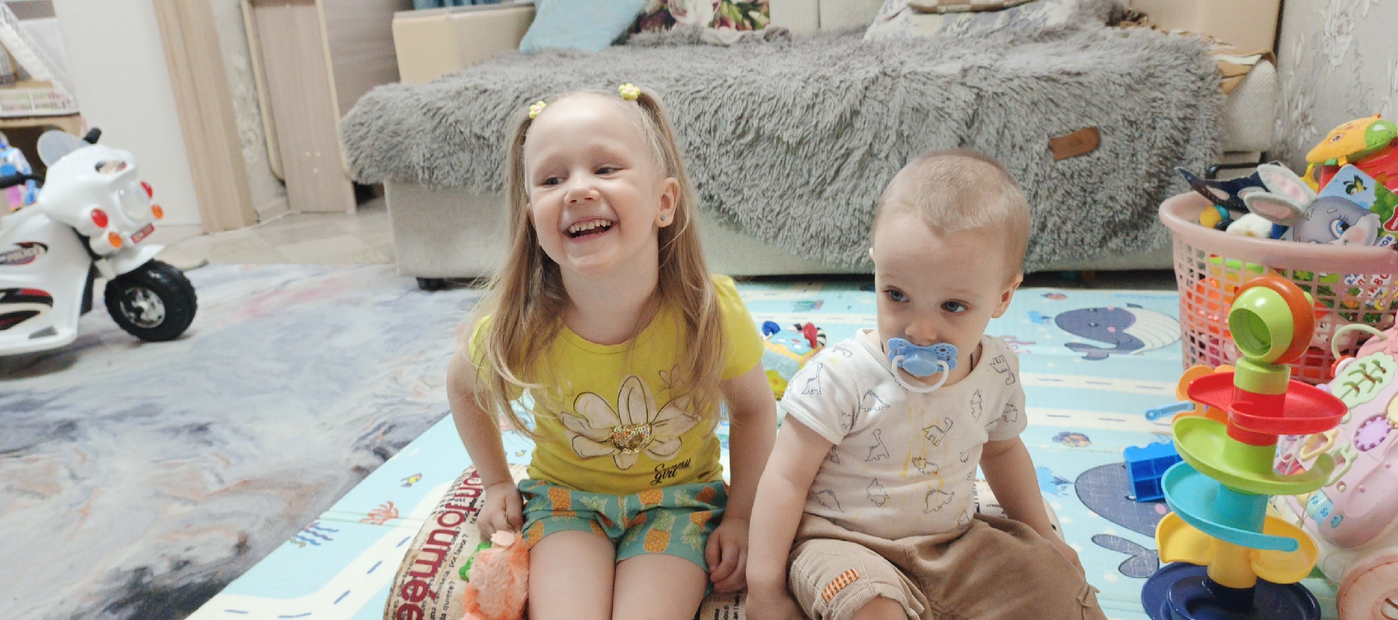 The image shows two young children sitting on a playmat in what appears to be a home in Chaikovsky, Russia. A fair-skinned girl with blonde hair pulled into pigtails, wearing a yellow T-shirt and pineapple-print shorts, is laughing. Beside her, a fair-skinned baby with a pacifier, wearing a dinosaur-print shirt and brown shorts, is seated. Toys are scattered around them, including a ball-drop toy and a basket of stuffed animals. A child's white motorcycle toy is visible in the background, along with a gray sofa and a light-colored wall. The playmat has a light blue background with cartoon images of whales, cars and roads.