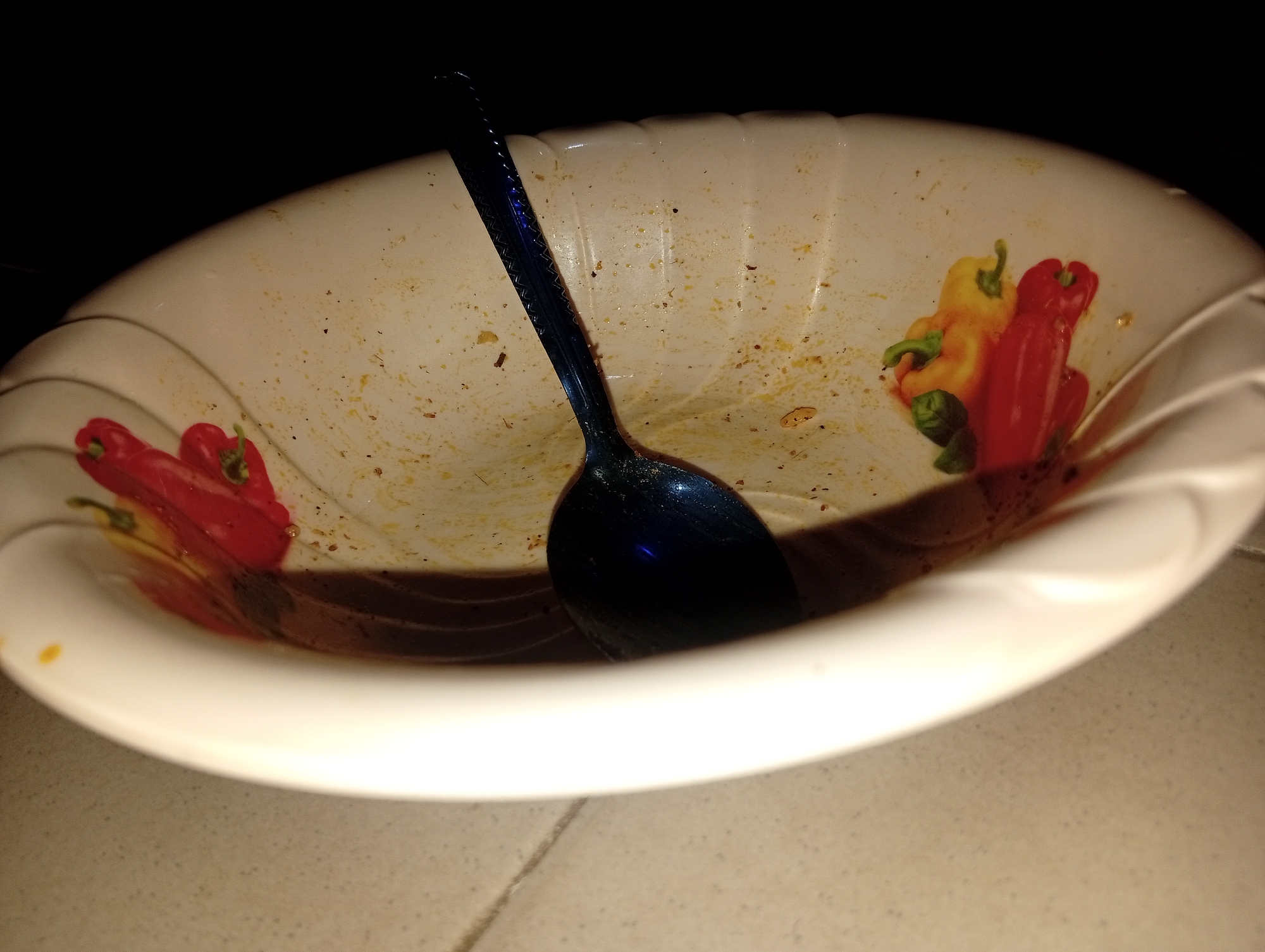 The image shows a white bowl with a decorative pattern of red and yellow bell peppers. The bowl contains some remaining liquid and has a dark blue spoon inside. The bowl is placed on a tiled surface. The background is dark. The scene appears to be an indoor domestic setting. There is no information available about the city of Ukukwa, Nigeria in relation to this image.