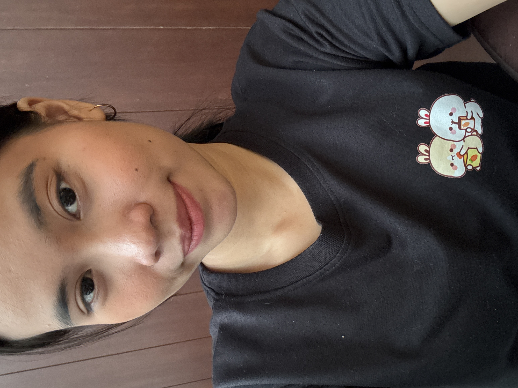 A young woman with dark hair and a black t-shirt with a cute bunny graphic is shown in a close-up selfie. Her face is turned towards the camera, and she is smiling slightly. The background appears to be a wooden wall. The image was taken in South Jakarta, Indonesia.