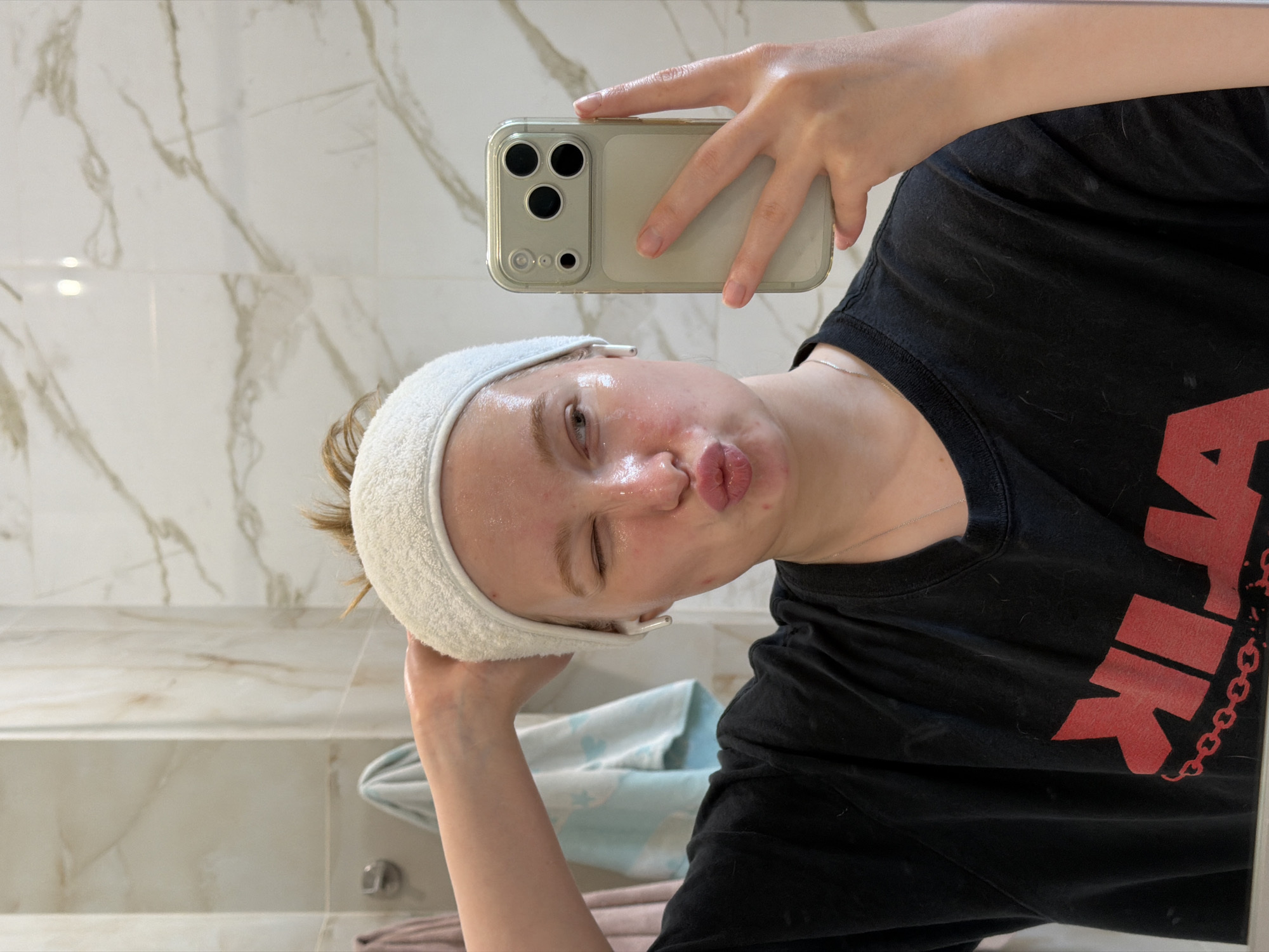 A young individual, appearing female, with reddish-blonde hair secured by a white terrycloth headband, is captured in a mirror selfie. Her face is visibly shiny, consistent with the application of a skincare product. She is winking with her right eye (viewer's left) and puckering her lips in a kissing gesture, while looking towards the right. White wireless in-ear earbuds are present in both ears. Her left hand is raised to touch the headband on the left side of her head, while her right hand holds a light-colored smartphone with a clear case, reflecting the mirror. The smartphone's rear camera array, featuring three large lenses and a smaller sensor, is clearly visible. She is wearing a black t-shirt with a bold red graphic, which includes a chain link pattern, on the chest. A thin silver-colored chain necklace is visible around her neck.
The background, visible in the mirror's reflection, consists of a bathroom interior with light-colored, possibly beige or white, tiled walls featuring a subtle marble-like veining pattern. Two towels, one light pink and one light blue with a white pattern, hang on hooks in the background. The specific location, including city and country, is not discernible from the visual information provided.