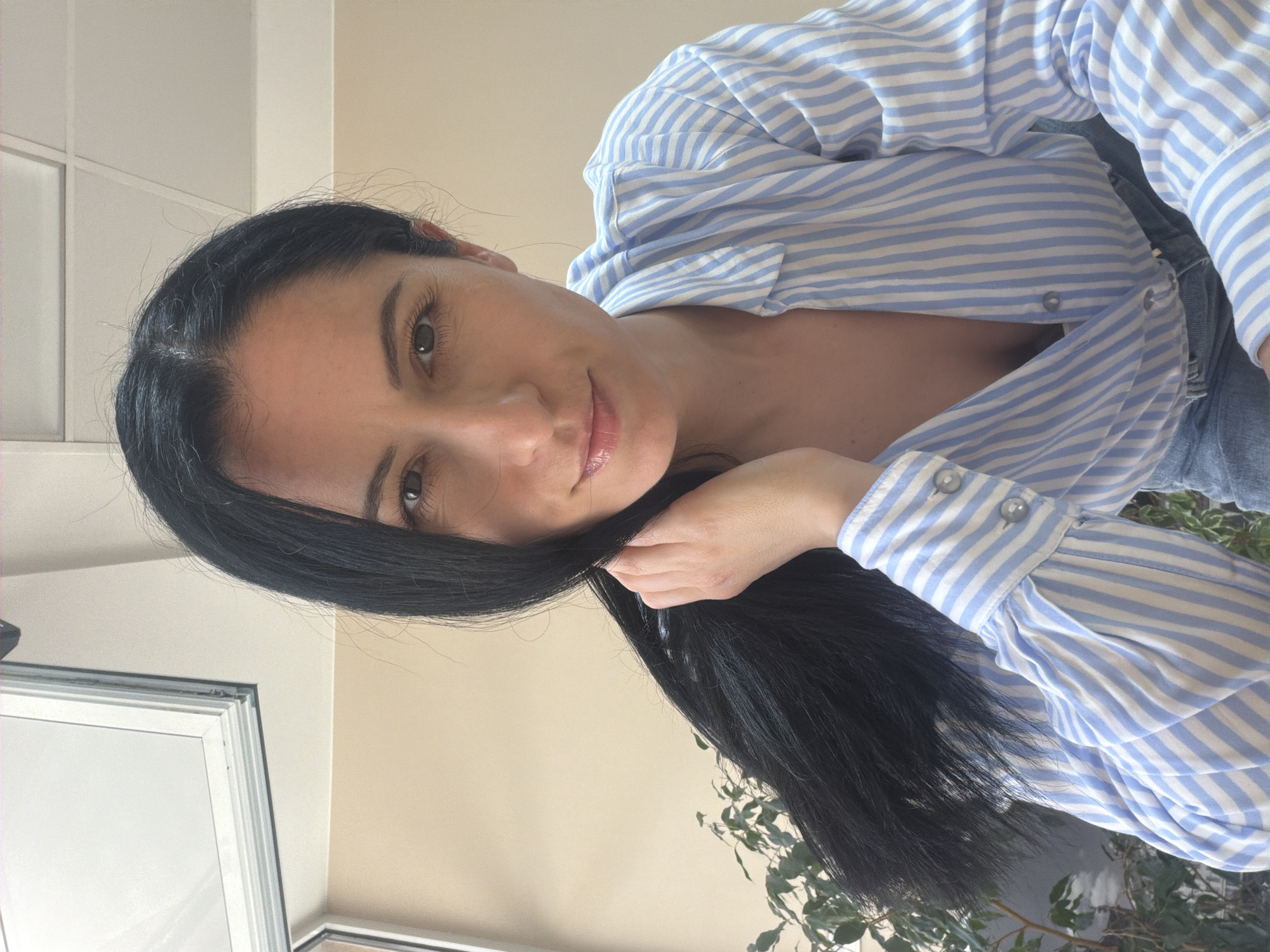 A woman with dark hair, wearing a blue and white striped shirt, is indoors. Her right hand holds a strand of her hair. The background includes a window and a plant. The location is unknown and cannot be determined from this image.