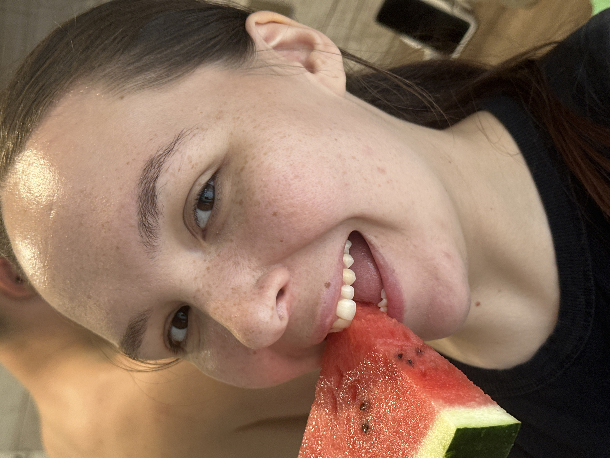 A close-up, slightly angled shot shows a young woman taking a bite out of a slice of watermelon. Her face is tilted towards the camera, with her mouth open as she bites into the fruit, revealing her teeth and tongue. She has numerous freckles across her face and a glistening sheen on her forehead, suggesting either sweat or makeup. Her eyebrows are dark and well-defined, and her eye, looking into the camera, reflects a bright light. The watermelon slice is a vibrant red with visible black seeds and a green rind. The background is blurred, showing hints of a person's bare shoulder on the left and a dark fabric on the right, possibly a shirt or sweater. There is also a dark rectangular object visible in the upper background, potentially a phone. The overall lighting suggests it could be daytime, possibly outdoors given the sheen on her skin. The image conveys a sense of enjoyment and refreshment associated with eating watermelon. There is no visible text in the image.