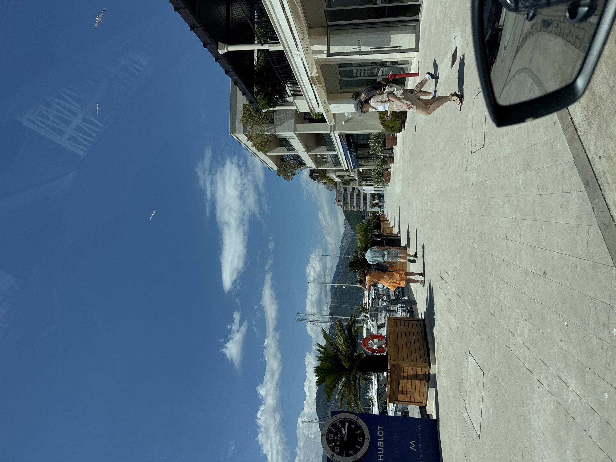 The image depicts a sunny day in Tivat, Montenegro, likely in Porto Montenegro, a luxury marina. The scene is a pedestrian walkway alongside a marina with boats docked. In the foreground, a woman wearing a light-colored outfit and a hat is walking, while a man walks behind her. To the left, a large Hublot clock is visible on a dark blue sign, with a palm tree and a wooden planter box nearby. Several people are walking along the pathway, and there are buildings with balconies and shops lining the right side of the walkway, one of which has a sign that reads "heidi klein." The sky is blue with scattered clouds, and seagulls are flying overhead. The perspective suggests the photo was taken from a vehicle, as a car's side mirror is visible in the lower right corner.