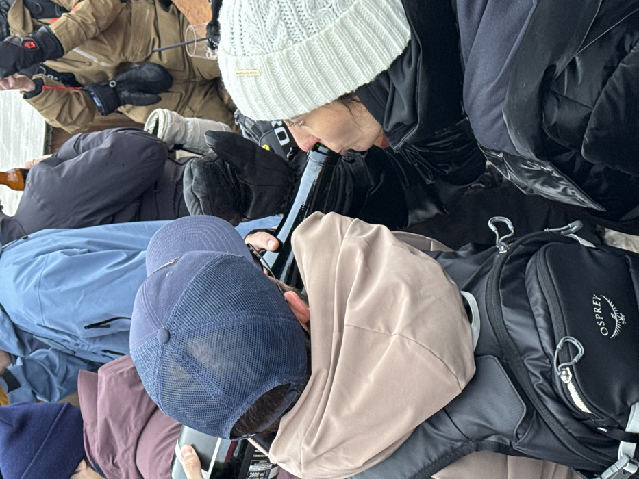 The image captures a close-up of several people seated closely together, possibly in an enclosed space. A woman in a white knit hat appears to be drinking from a dark glass bottle. Nearby, an individual wears a blue baseball cap and another has a tan-colored hood pulled up. An Osprey backpack is visible, as are various winter coats and gloves, suggesting a cold environment. A glass with what appears to be a beverage can be seen among the garments.