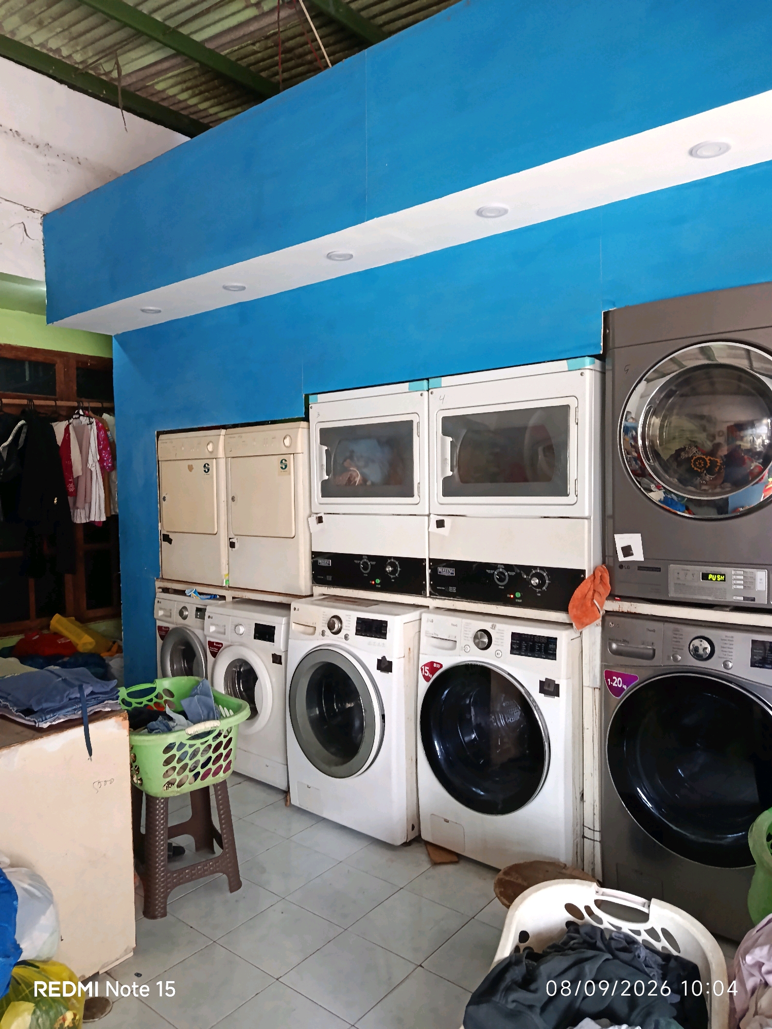 The image depicts the interior of a laundromat in Keboansikep, Indonesia, with multiple washing machines and dryers arranged in rows. The space is characterized by a bright blue wall with a white stripe running horizontally across it, installed with recessed lighting. In the foreground, a green laundry basket filled with clothes sits atop a brown stool. To the left, a portion of a beige counter is visible, with some laundry supplies and a blue garment on top. The floor is tiled with a pattern of light and dark gray squares. Clothing is also visible hanging on a rack to the left, suggesting ongoing laundry services. The overall scene is functional and appears to be in operation, with the presence of various appliances indicating a commercial establishment. The timestamp "08/09/2026 10:04" is overlaid on the bottom right of the image, suggesting it was taken on that date and time.