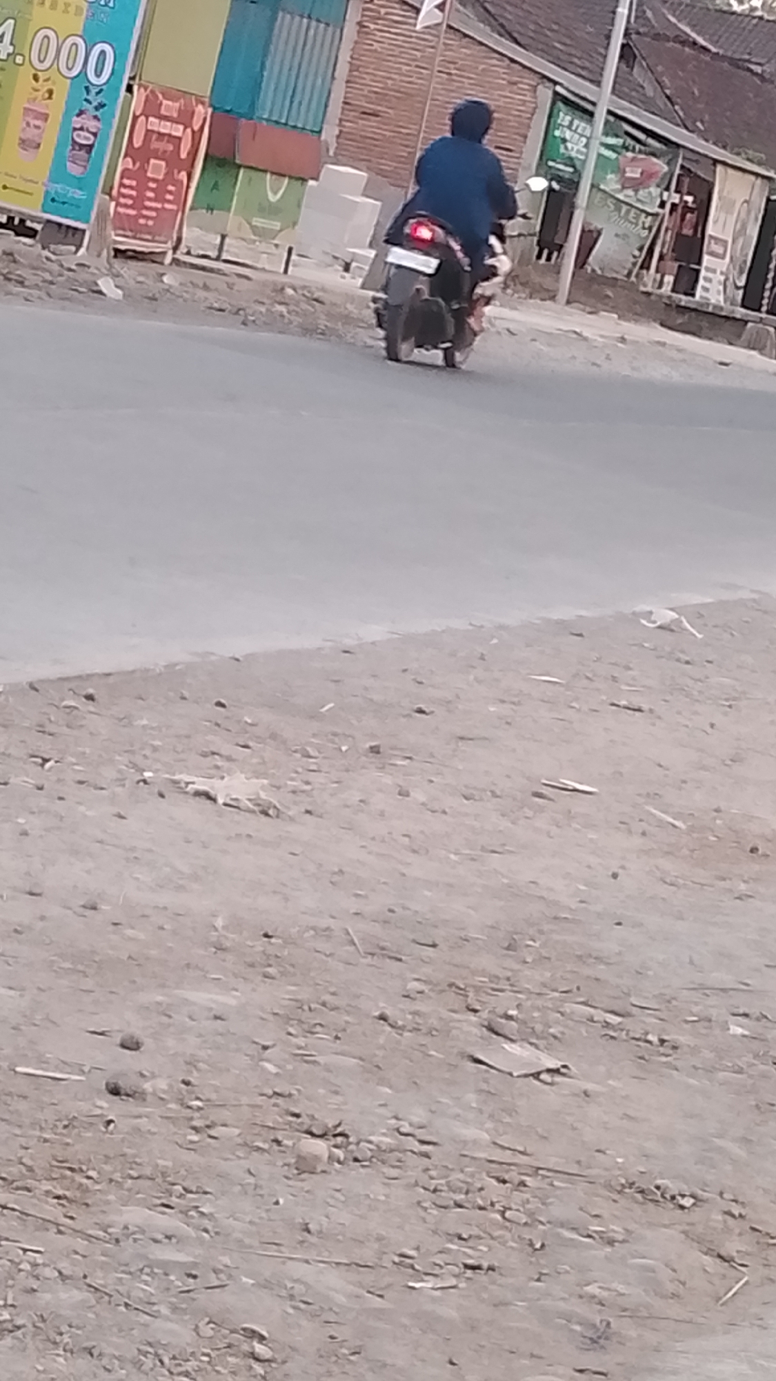 A lone motorcycle rider, wearing a dark blue jacket and helmet, is captured from behind as they drive away on a paved road. The rider is on a dark-colored motorcycle with visible taillights. The scene appears to be in a semi-urban or rural area, possibly a roadside in Karanglangu, Indonesia, given the presence of a building with red brick walls in the background and advertisements on a stand to the left, one of which displays "4.000". The ground in the foreground is unpaved, with scattered debris and small stones, suggesting a less developed or maintained area alongside the road. The lighting indicates it is daytime, with the sun casting some shadows.