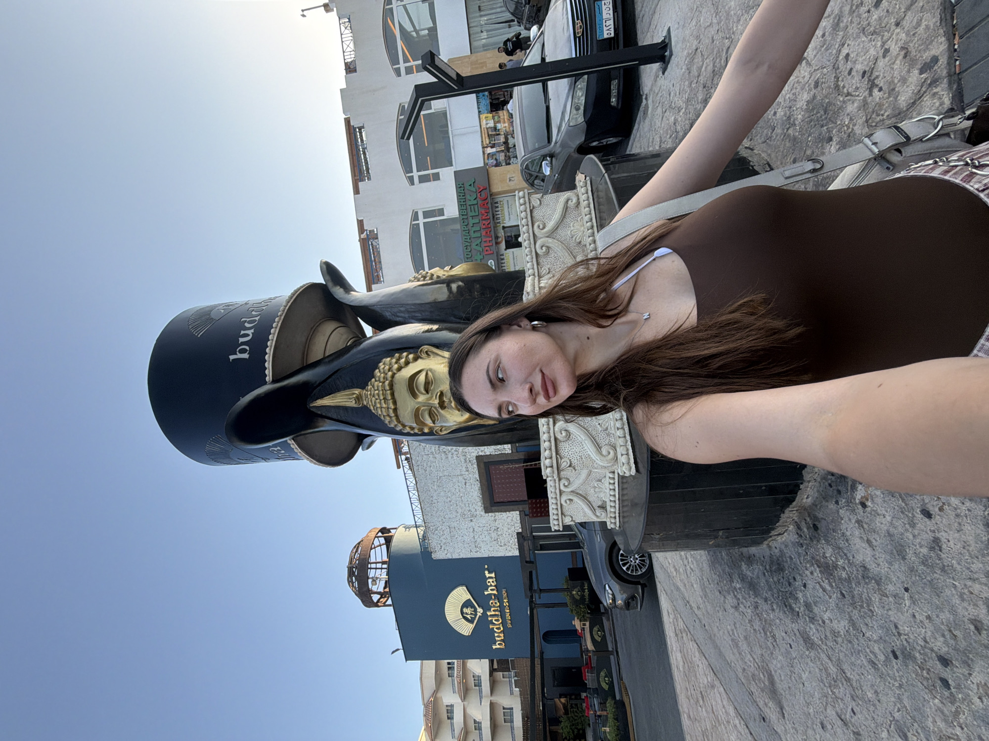 A young woman with long brown hair is taking a selfie in front of a large sculpture of Buddha heads. She is wearing a brown tank top and a silver necklace with a pendant shaped like the letter "N". Behind her, a building with the "Buddha-Bar Sharm El Sheikh" logo is visible. Further in the background, a pharmacy with Russian signage is located on a white building. Several cars are parked along the street, and the sky is clear and blue. The scene appears to be an outdoor public space in Sharm El Sheikh, Egypt.