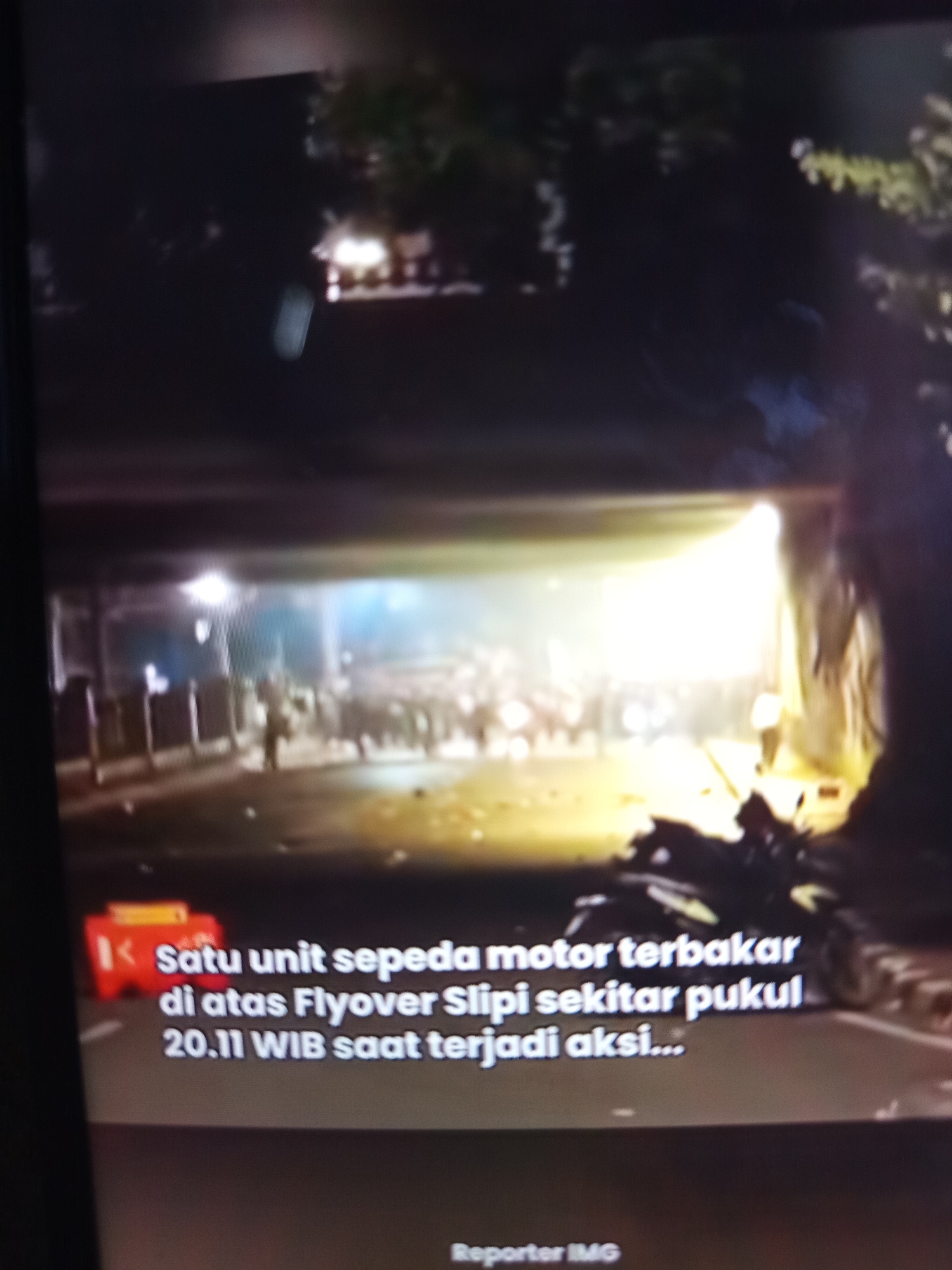 The image depicts a dark, nighttime scene with a bright light illuminating a street under an overpass. There appears to be a gathering of people, and in the foreground, a portion of text overlay is visible on a red and yellow graphic. The text reads "Satu unit sepeda motor terbakar di atas Flyover Slipi sekitar pukul 20.11 WIB saat terjadi aksi..." which translates to "One motorcycle burned on the Slipi Flyover around 20.11 WIB when an action occurred...". The scene suggests a chaotic event, possibly a protest or riot, involving a burning motorcycle and a crowd. The location is identified as Flyover Slipi, which is in Jakarta, Indonesia, not Ngablak.