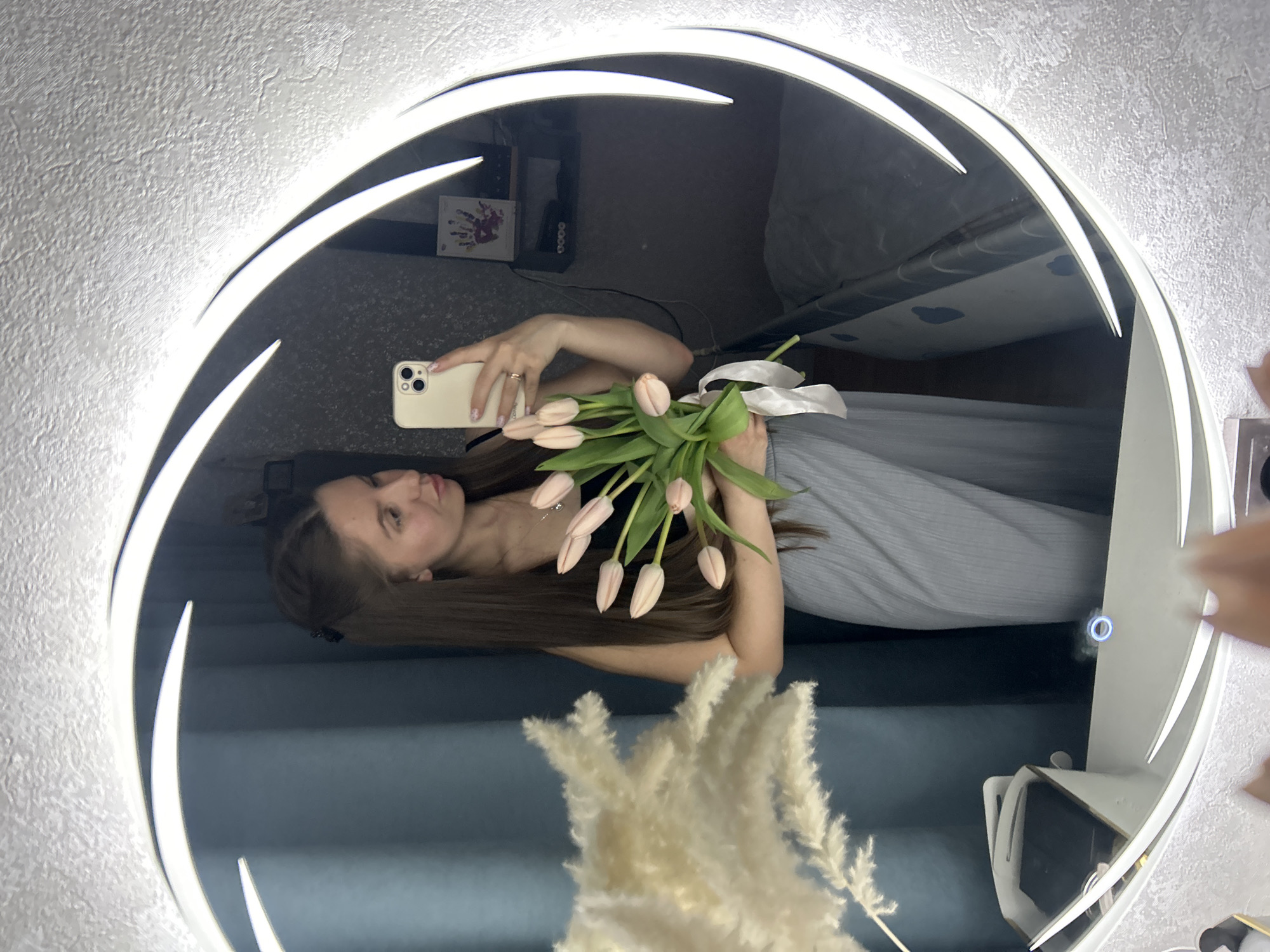 A young woman with long brown hair stands in front of a round mirror with a bright, white LED ring, taking a selfie with a beige smartphone. She is holding a bouquet of pink tulips tied with a white ribbon. The reflection shows her wearing a gray dress with vertical stripes. The background includes a dark shelf with objects, a gray bed with a patterned sheet, and a blue wall. A vase with dried pampas grass is partially visible in the foreground. The location is not explicitly identifiable as Klin, Russia, from the visual content of the image.