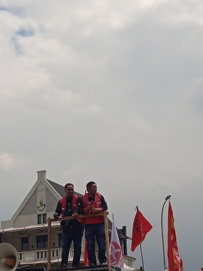 Two men, both wearing red vests and red headbands, are standing on a wooden platform in front of a white building. The building has a gable roof and a green sign on the side. The men are holding microphones and are looking out at the crowd. There are two red flags in the background, one behind the men and one to the right. There is also a white flag with a red symbol on it in the foreground. The sky is cloudy. The location is at Jalan Jendral Sudirman, Yogyakarta, Indonesia (latitude: -7.783, longitude: 110.367). A white loudspeaker is visible on the left side of the frame. The men are speaking into the microphone.