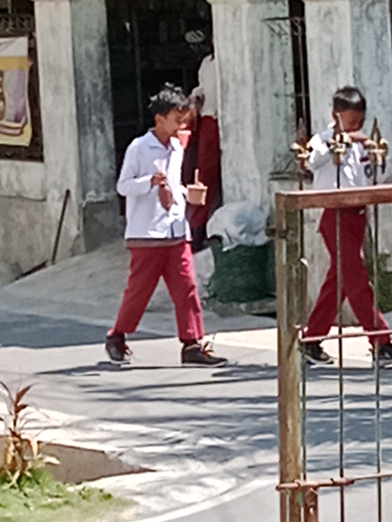 The image, taken in Waru, Indonesia, depicts two young individuals in what appears to be a school uniform. The individual on the left is wearing a white long-sleeved shirt and red trousers. They are walking away from the camera and holding a light-colored object in their right hand. Their left hand is by their side. The individual on the right is also wearing a white shirt and red trousers, and is walking towards the right side of the frame, away from the viewer. They are holding something in their right hand and their left hand is extended forward. The scene appears to be outdoors, with a concrete pathway or road in the foreground and a building with a grey concrete wall in the background. There is also a metal fence visible on the right side of the image. The lighting suggests it is daytime, with shadows cast on the ground.