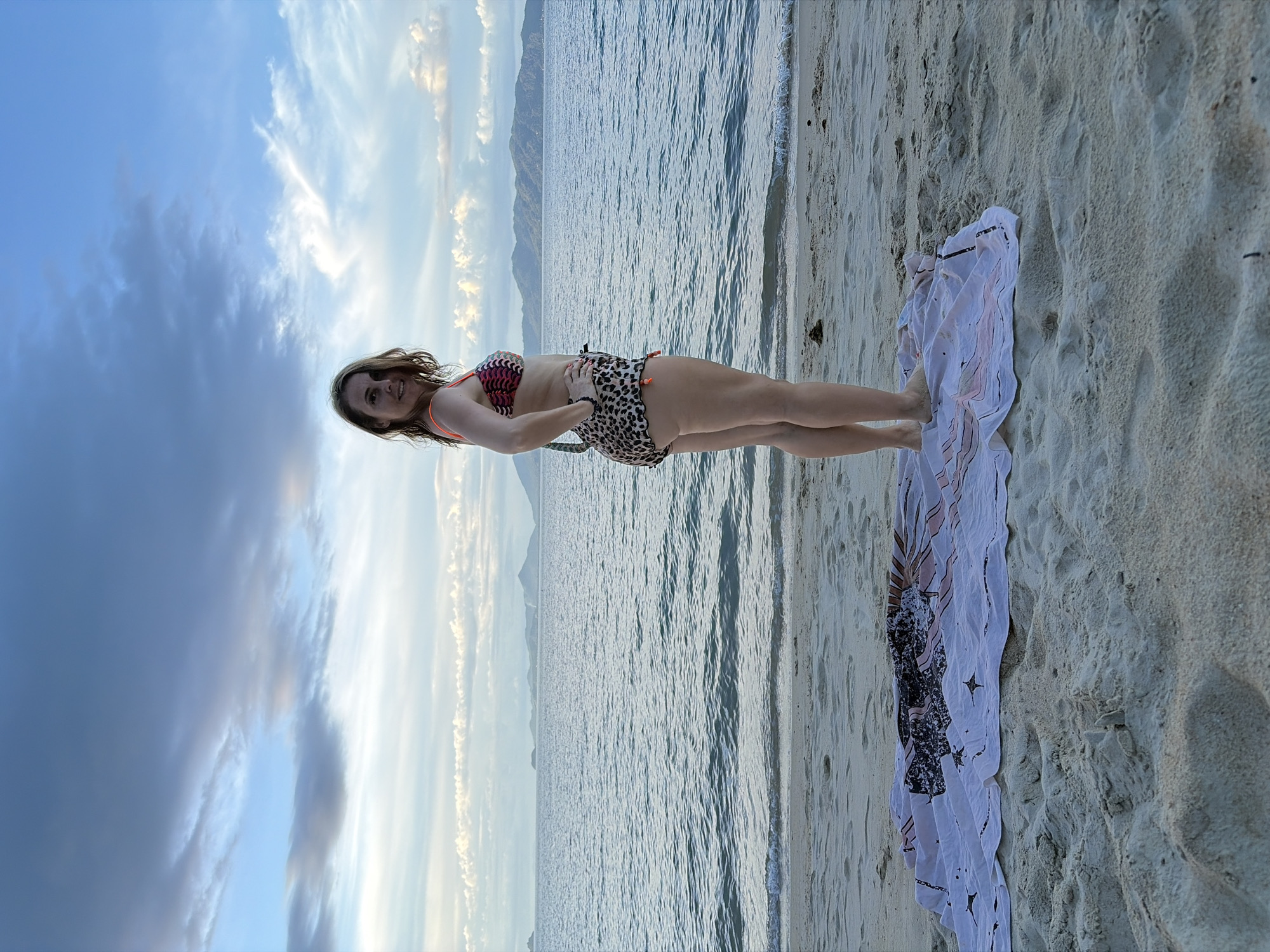A woman stands on a sandy beach in Baan Mae Nam, Thailand, at dusk. She is wearing a bikini with a leopard print bottom and a patterned top with orange straps. She is facing away from the camera, looking over her shoulder with a smile. In front of her, a white beach towel with a pink and black sunburst pattern is spread on the sand. The sea is calm with small waves lapping at the shore, and islands and mountains are visible in the distance under a cloudy sky.