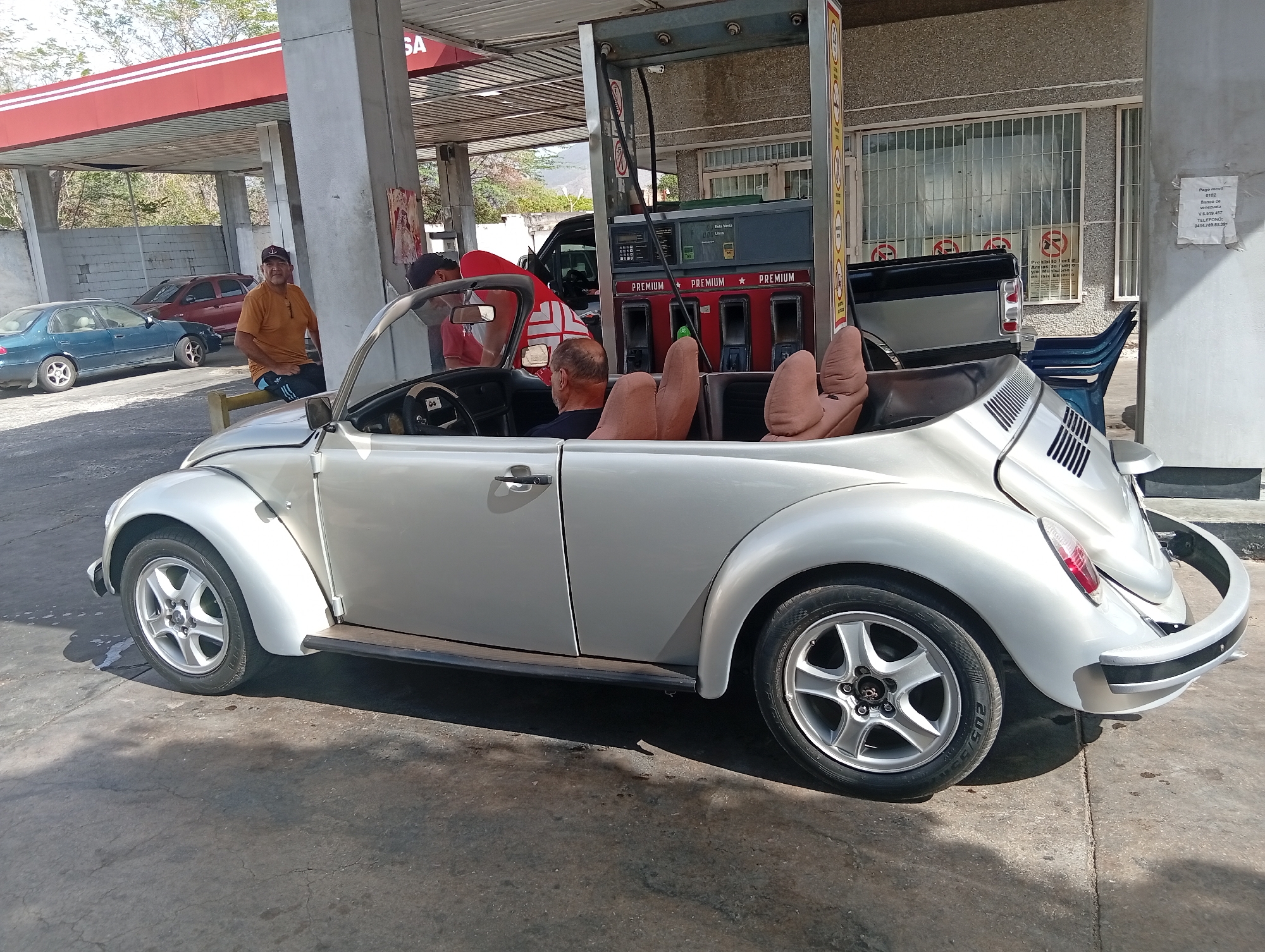 The image shows a silver Volkswagen Beetle convertible at a "PREMIUM" gas pump at a "SA" gas station. A man is sitting in the driver's seat of the car, and two strange looking seatbacks made from stuffed material are behind. Another man in an orange shirt is seated nearby, leaning against a barrier. A truck is being refueled at an adjacent pump. In the background, other vehicles are parked. A sign with "Pago movil" and a telephone number is attached to a pillar. The text on the pump also indicates that this is in Venezuela. Based on the OCR, this is in La Tagua, Venezuela.