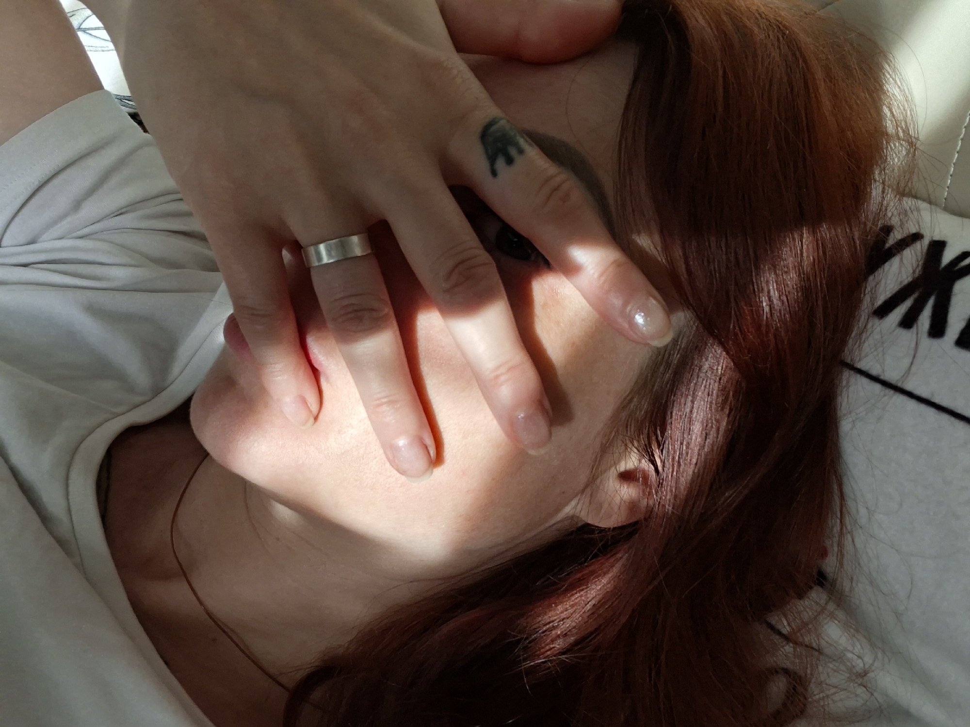 A close-up shot captures a person's face partially covered by their hand. The individual has long, reddish-brown hair and is wearing a white t-shirt. A silver ring is visible on one finger, and a small crown tattoo is on another. The lighting suggests the sun is casting shadows across the face, creating a dramatic effect. The background appears to be a light-colored surface, possibly a bed or couch. The image is taken indoors, and there are no discernible elements that would indicate a specific location within the city of Myadzyel, Belarus.