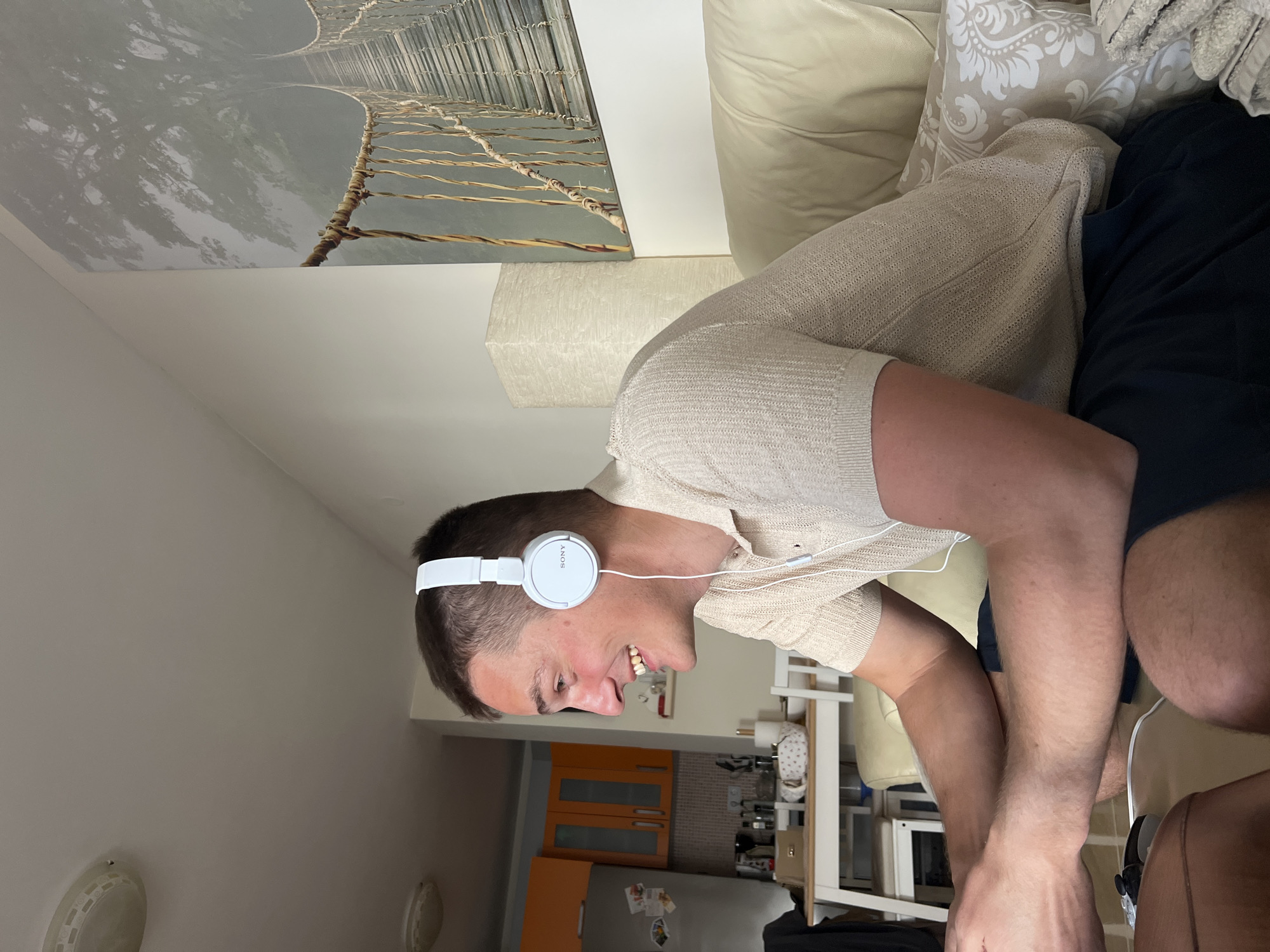A person is wearing white Sony headphones while sitting indoors on a light-colored couch. The person is wearing a tan shirt and dark shorts. Behind the person, there is a kitchen area with orange cabinets and a stainless steel refrigerator. Above the couch, there is a picture of a rope bridge, possibly a wallpaper. The exact location in Budva, Montenegro, cannot be determined from this image.