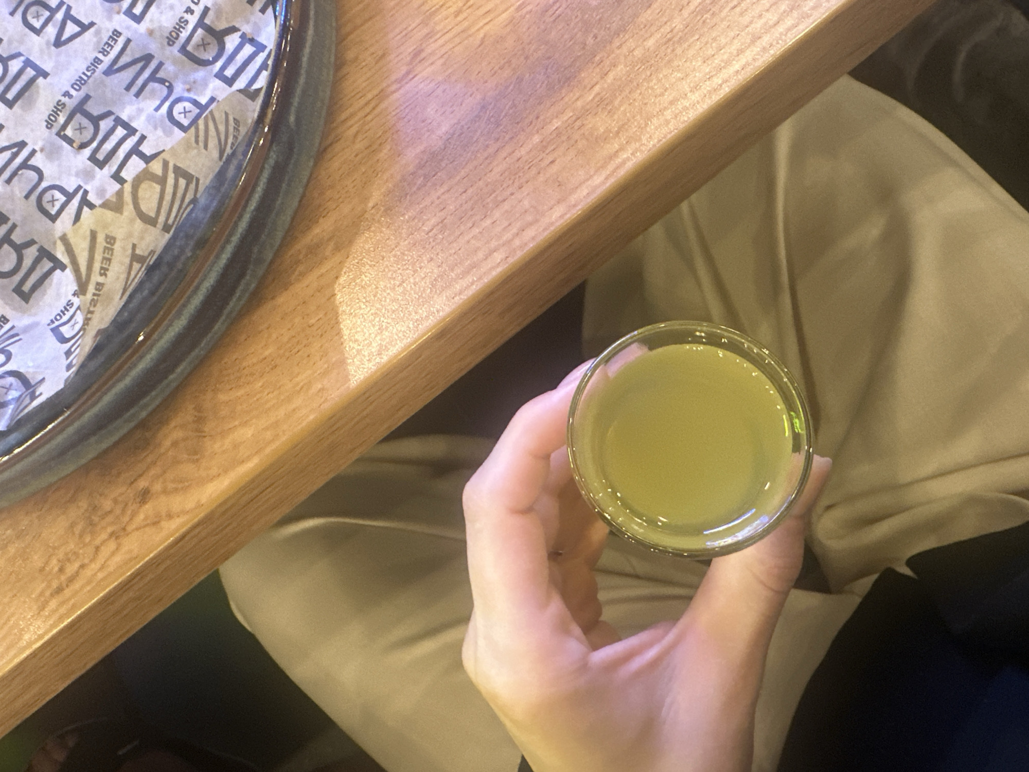 A high-angle shot captures a person holding a glass filled with a light yellow liquid, likely a beverage, at a table in what appears to be a restaurant or bar setting. The table, made of light-colored wood, is partially occupied by a plate with printed text reading "BEER BISTRO & SHOP." The person holding the glass is wearing light-colored clothing, and a dark garment is visible near their arm. This image was taken in the city of Perm, Russia.