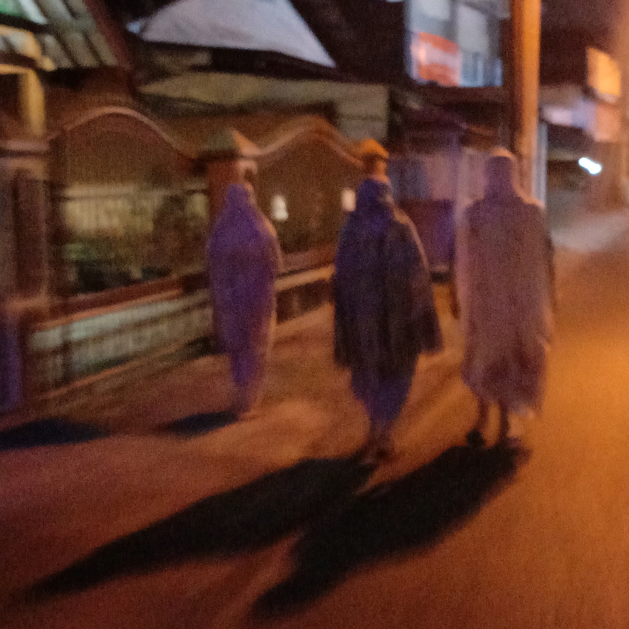 Three figures, silhouetted and blurred by the low light and possibly motion, walk along a street at night. The figures are dressed in dark, flowing garments that obscure their features. The first figure on the left is wearing a purple garment. The middle figure appears to be wearing a dark blue or purple garment and a bright yellow head covering. The figure on the right is wearing a lighter-colored garment, possibly white or light grey. Their shadows are cast long and distorted on the asphalt road, suggesting a low light source, likely a streetlamp or artificial lighting from nearby buildings.
The background shows the facade of buildings with illuminated windows and architectural details. A streetlamp or light pole stands prominently on the right side of the image. The environment suggests a residential or commercial street in an urban setting. Given the prompt's context, this scene is observed in Salatiga, Indonesia, at night. The overall impression is one of movement and anonymity in the dim, atmospheric glow of the city at night.