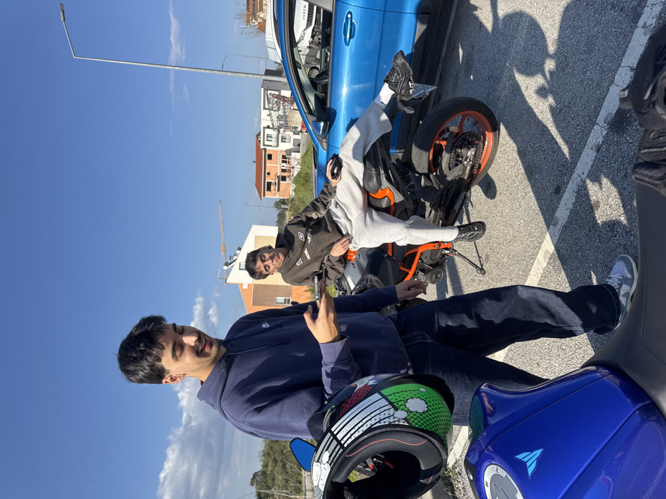 This image captures two young adult males and three vehicles in an outdoor parking area in Mirandela, Portugal, under a clear blue sky with minimal cloud cover.
The male on the left, wearing a dark blue Nike hooded sweatshirt, dark jeans, and light-colored sneakers, stands holding a smartphone in his left hand and a motorcycle helmet with green, white, and red graphic elements in his right hand. He is looking down at his phone with a slight smile. A blue Yamaha MT series motorcycle is partially visible in the immediate foreground, to his left.
The male on the right, wearing a dark brown hooded sweatshirt, light grey sweatpants, and dark athletic shoes, sits casually on an orange and black sports motorcycle, which is resting on its side stand. His left leg is extended forward, and his right leg is bent. He holds a small black object in his right hand and looks towards the camera with a smirk. Behind him, a blue SUV with an "IVECO" logo visible on its rear quarter is parked.
The ground consists of grey asphalt with faded white parking lines. In the background, there is sparse vegetation, an orange-brown residential building, and a multi-story building under construction with exposed concrete frames and red brickwork. A yellow construction crane is visible in the far distance, behind the buildings. A tall street light pole stands on the right side of the frame. Strong sunlight casts distinct shadows across the scene.