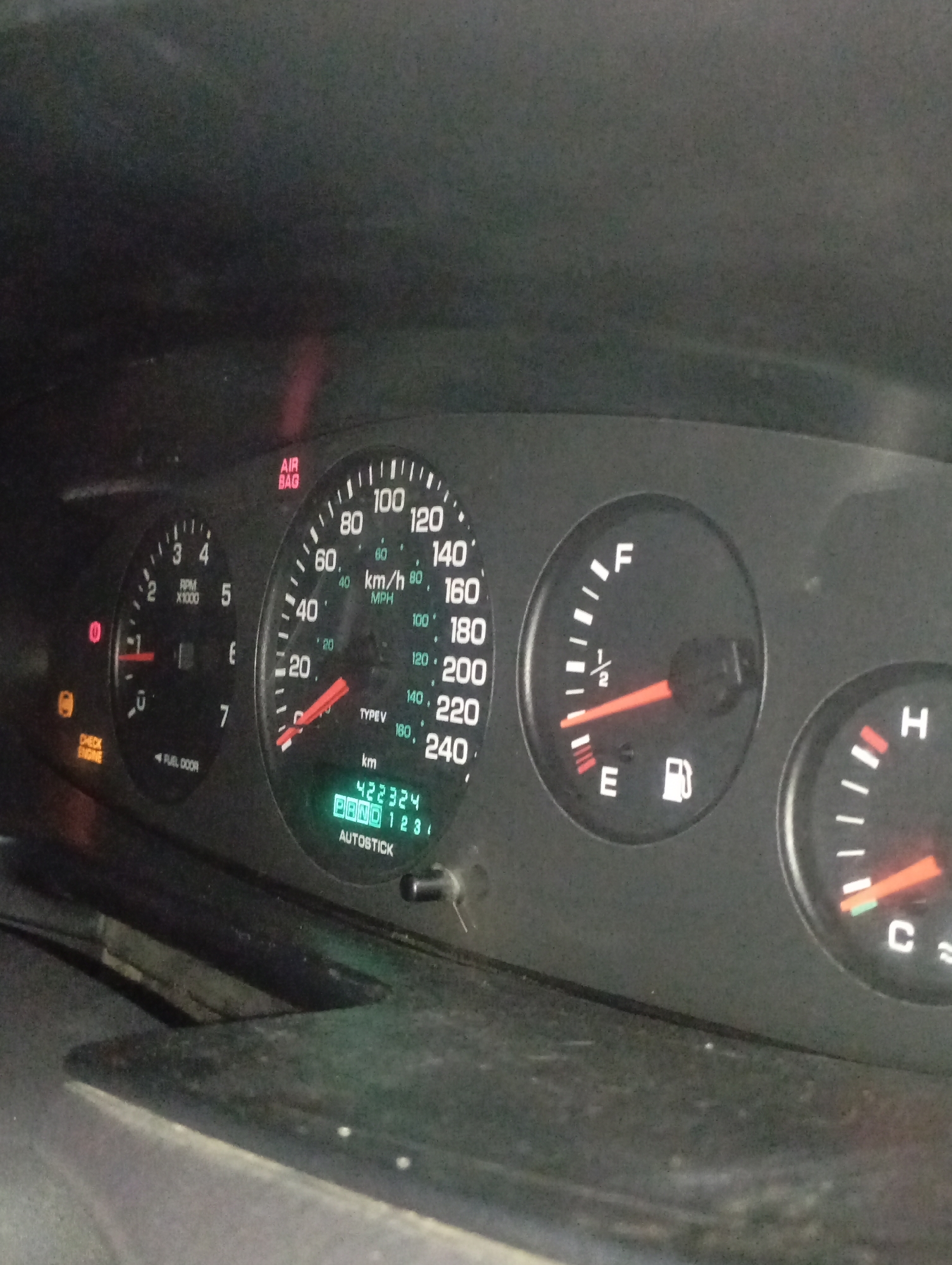 This image depicts an automobile dashboard instrument cluster, located within a vehicle in Kurganinsk, Russia. The scene is viewed from the driver's perspective, displaying a dark gray dashboard surface with visible dust. The speedometer, marked 0-240 km/h (0-150 mph), indicates a speed of approximately 20-30 km/h (15-20 mph). The tachometer shows a needle positioned just above 0 RPM. The fuel gauge needle rests near 'E' (Empty), with the fuel pump icon visible. The temperature gauge needle is at the 'C' (Cold) mark. Several warning indicators are illuminated: a red 'AIR BAG' light, an orange 'CHECK ENGINE' light, and a red circular light with an exclamation mark. The digital odometer displays '422324 km'. Below the odometer, 'P' (Park) is illuminated in green, alongside 'RND 1 2 3' and the text 'AUTOSTICK'. No individuals or specific actions are visible.